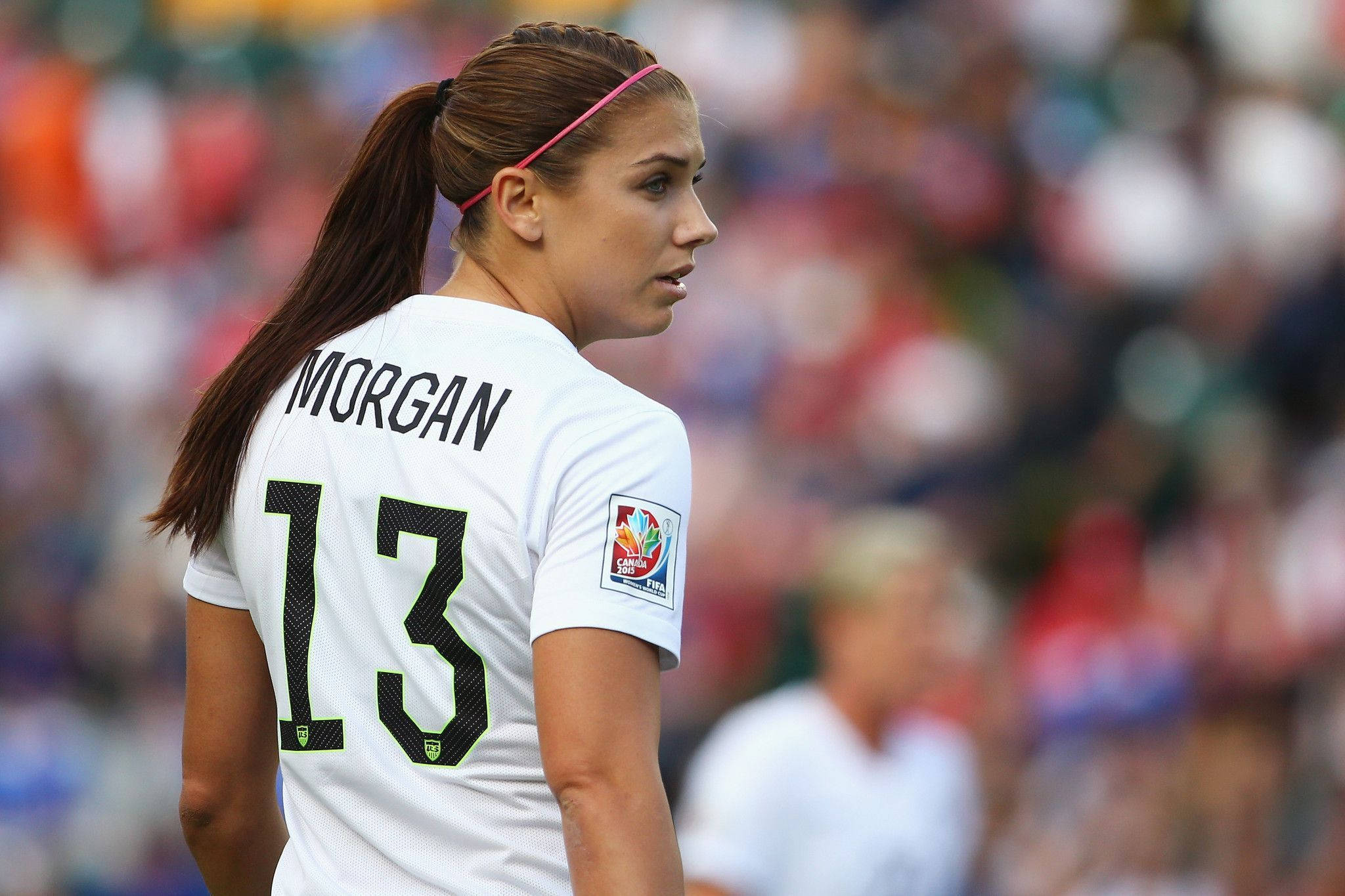 Alex Morgan 桌布 壁紙 — 一位專注的女足球員，身穿白色球衣，上面有名字 morgan 和號碼 13，站在陽光明媚的球場上，背景是模糊的觀眾。