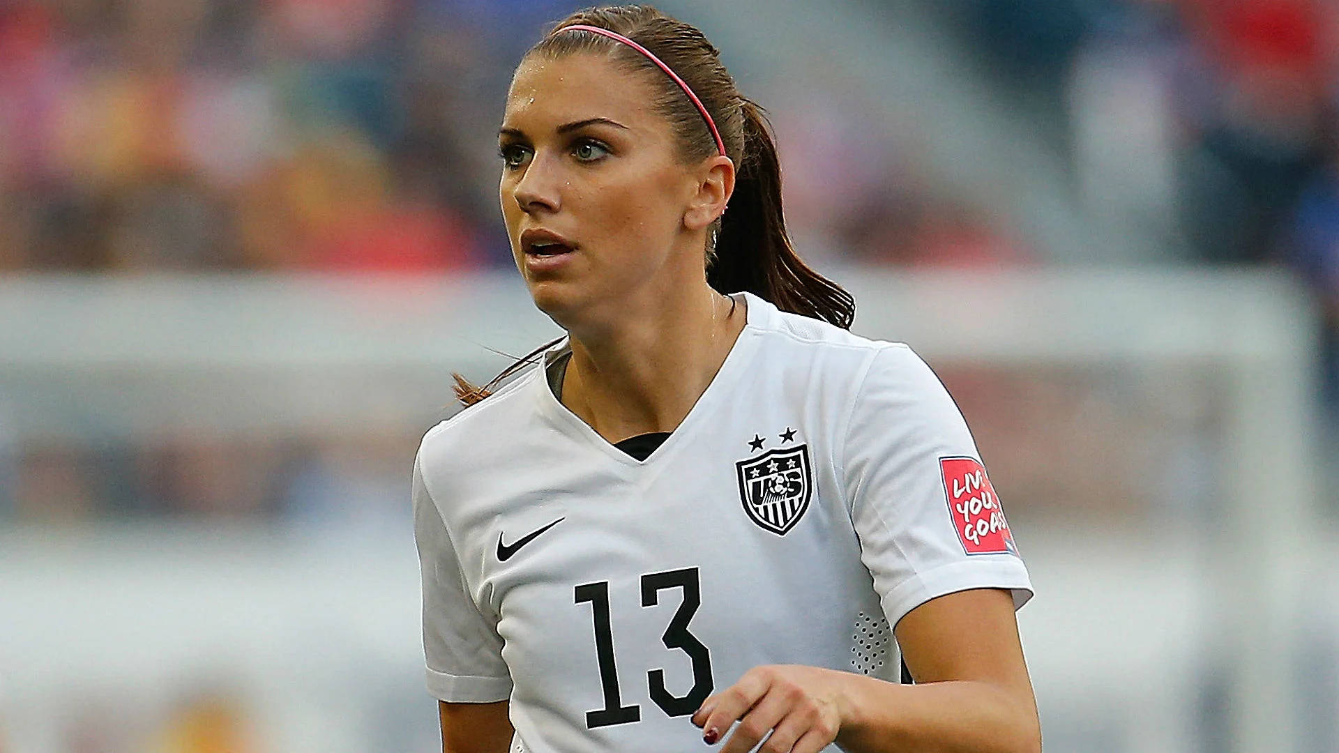 Alex Morgan 桌布 壁紙 — 一位專注的女足球員，身穿白色美國國家隊13號球衣，在日間比賽中奮力奔跑。