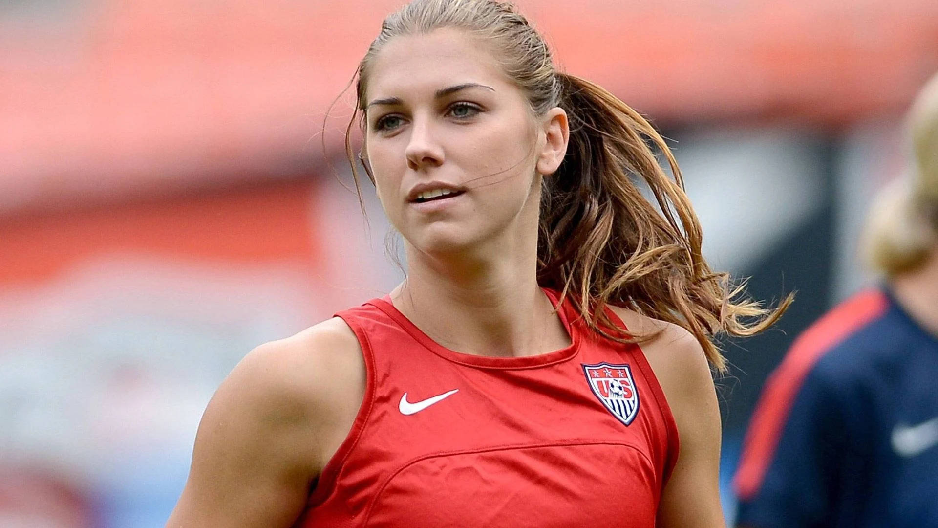 Alex Morgan 桌布 壁紙 — 一位專注的女足球員穿著紅色美國國家隊球衣，在日間訓練中決心奔跑。