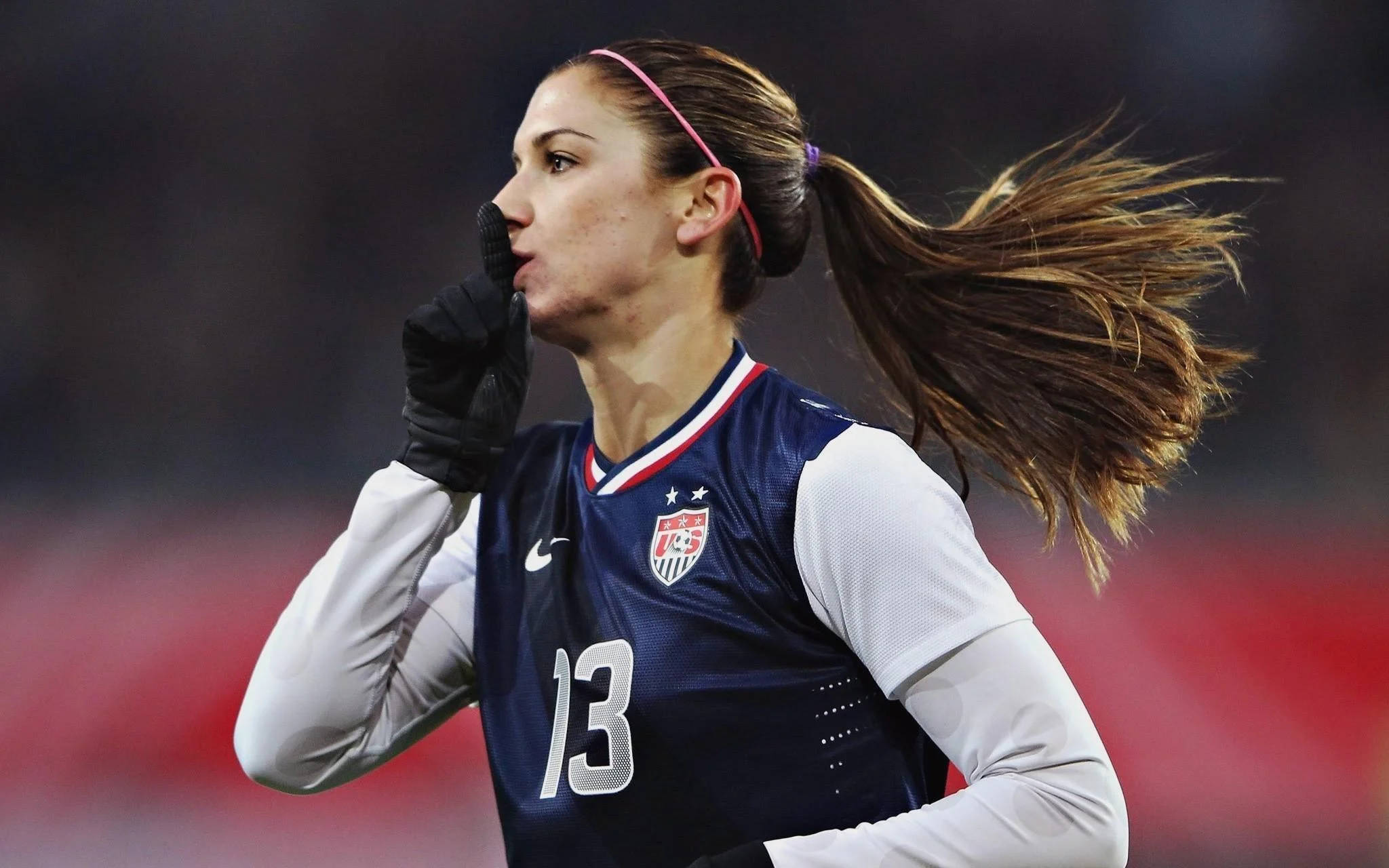 Alex Morgan 桌布 壁紙 — 一位專注的女足球員穿著美國球衣奔跑，馬尾辮飛揚，手靠近嘴巴。