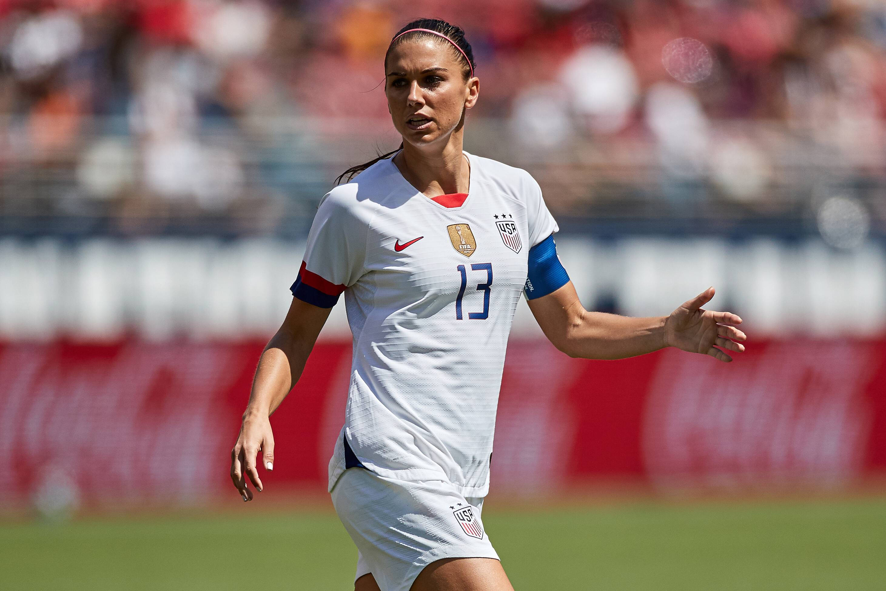Alex Morgan 桌布 壁紙 — 一位專注的女足球員身穿白色美國國家隊13號球衣，在充滿活力的綠色球場上進行日間比賽時做出手勢。