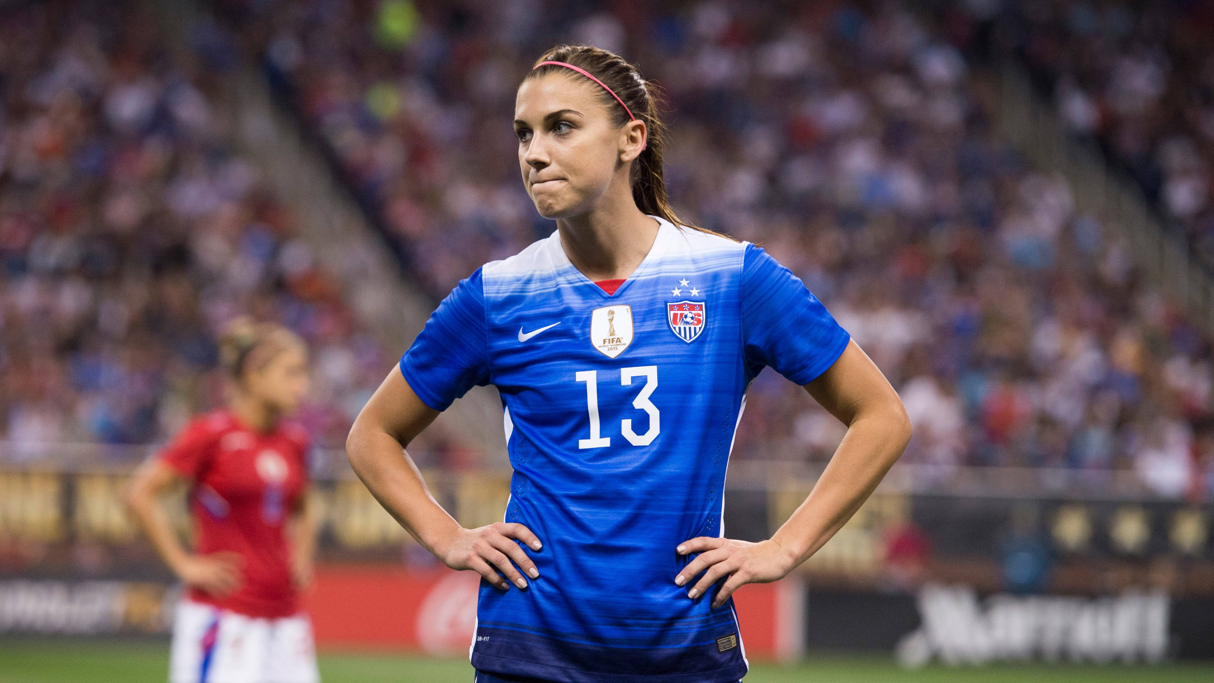 Alex Morgan 桌布 壁紙 — 一位專注的女足球員身穿藍色美國隊13號球衣，在比賽期間雙手叉腰站在體育場的球場上。