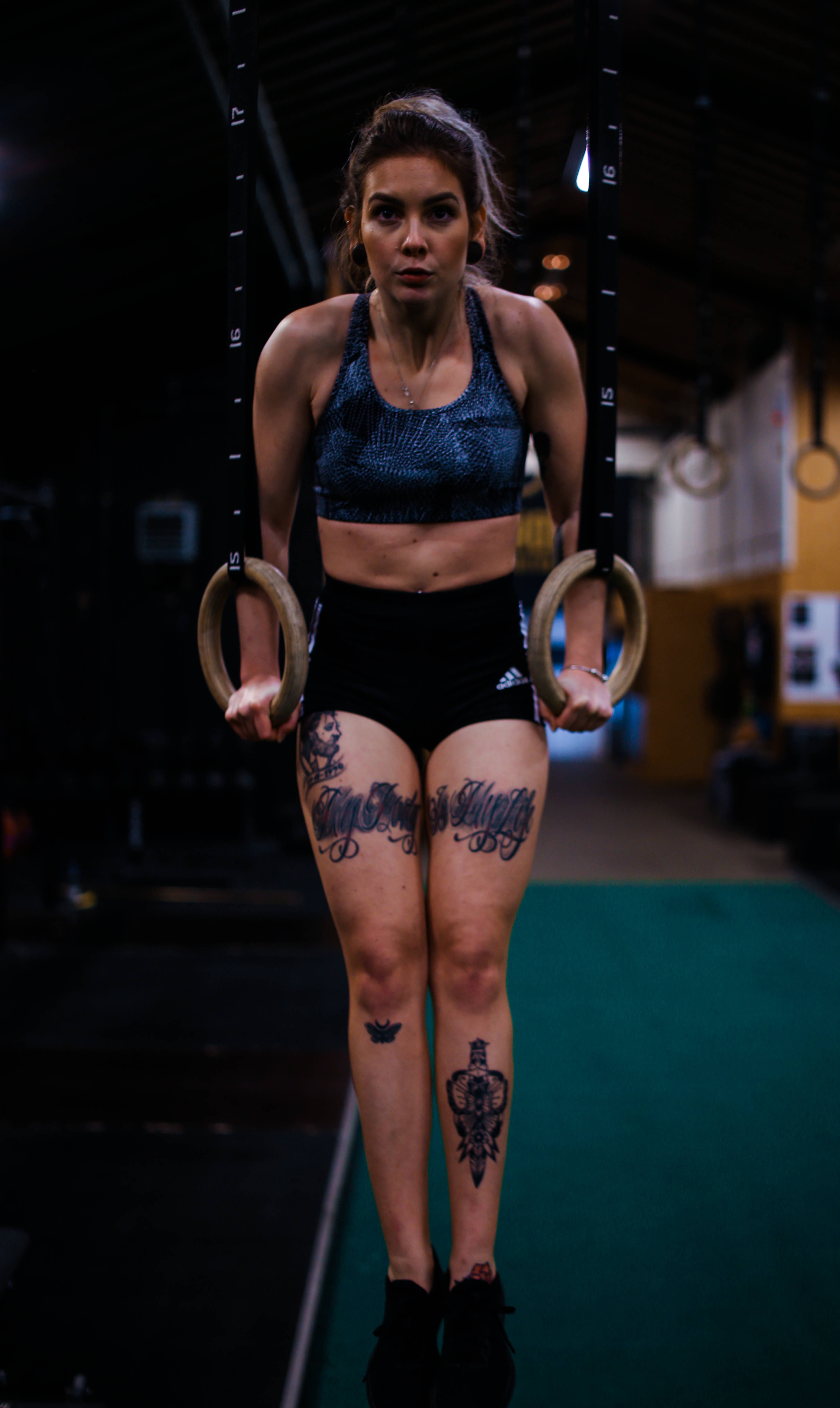 Athletic 桌布 壁紙 — 一位專注的女運動員懸掛在體操環上，在昏暗的健身房中，她腿上的刺青清晰可見。