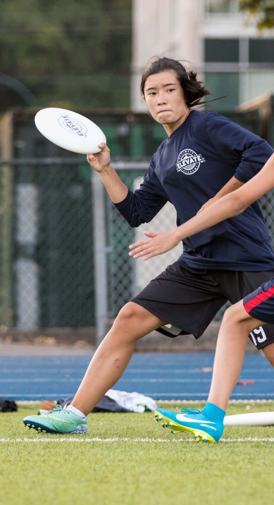 Athlete 桌布 壁紙 — 一位專注的女運動員穿著海軍藍襯衫和亮藍色鞋子，準備在跑道附近的草地上投擲白色飛盤。