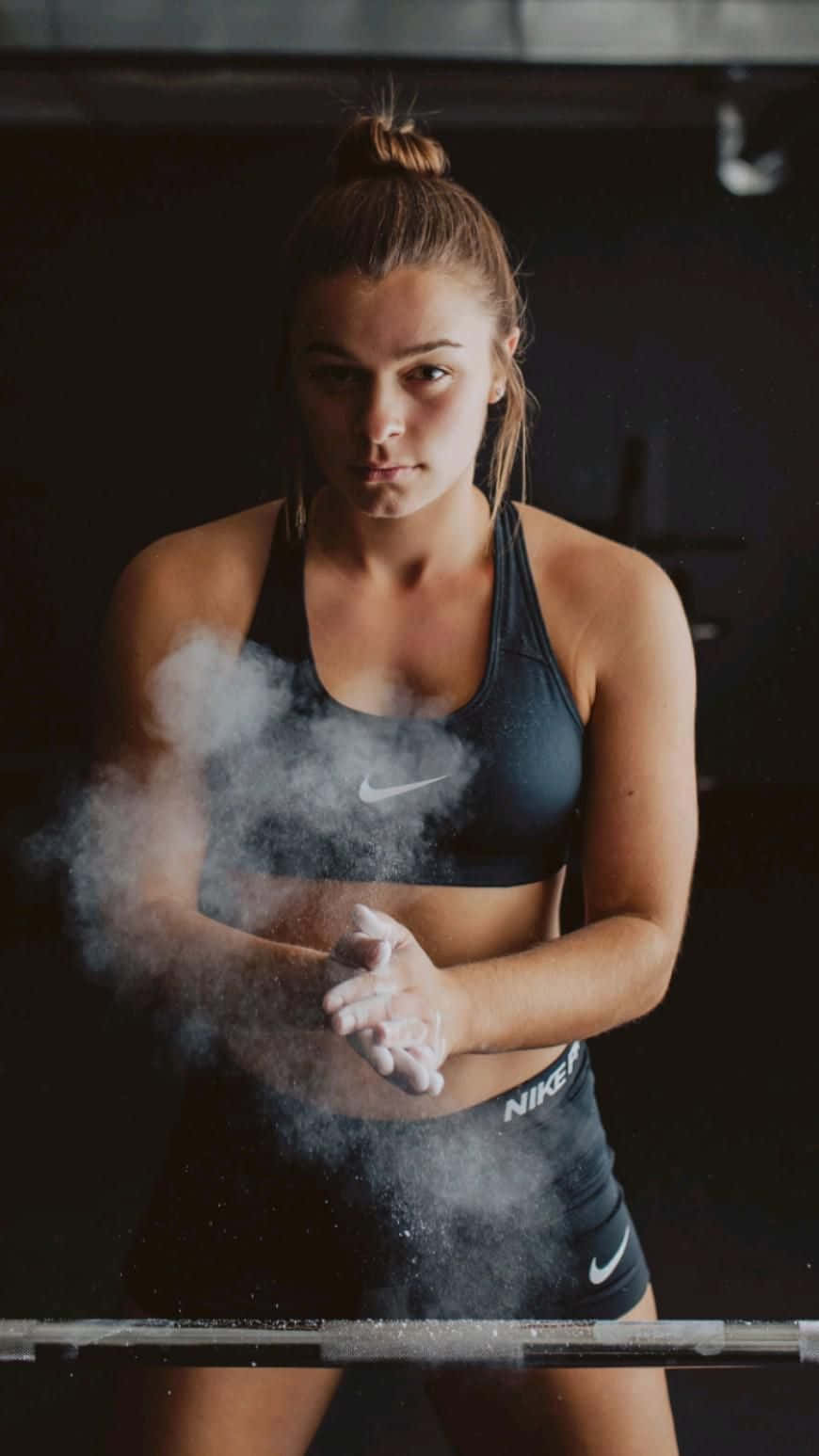 健身房 桌布 壁紙 — 一位專注的女運動員穿著深色Nike裝備，在昏暗的健身房中舉起槓鈴前，在手上塗抹粉筆灰。