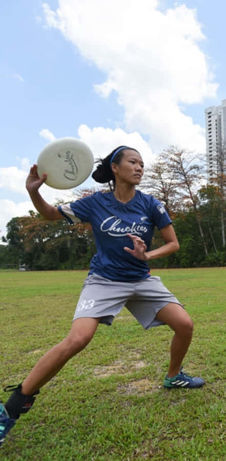 Android Ultimate Frisbee 桌布 壁紙 — 一位專注的女運動員穿著藍色球衣，在陽光明媚的草地上擲出白色飛盤，背景有樹木和高樓大廈。