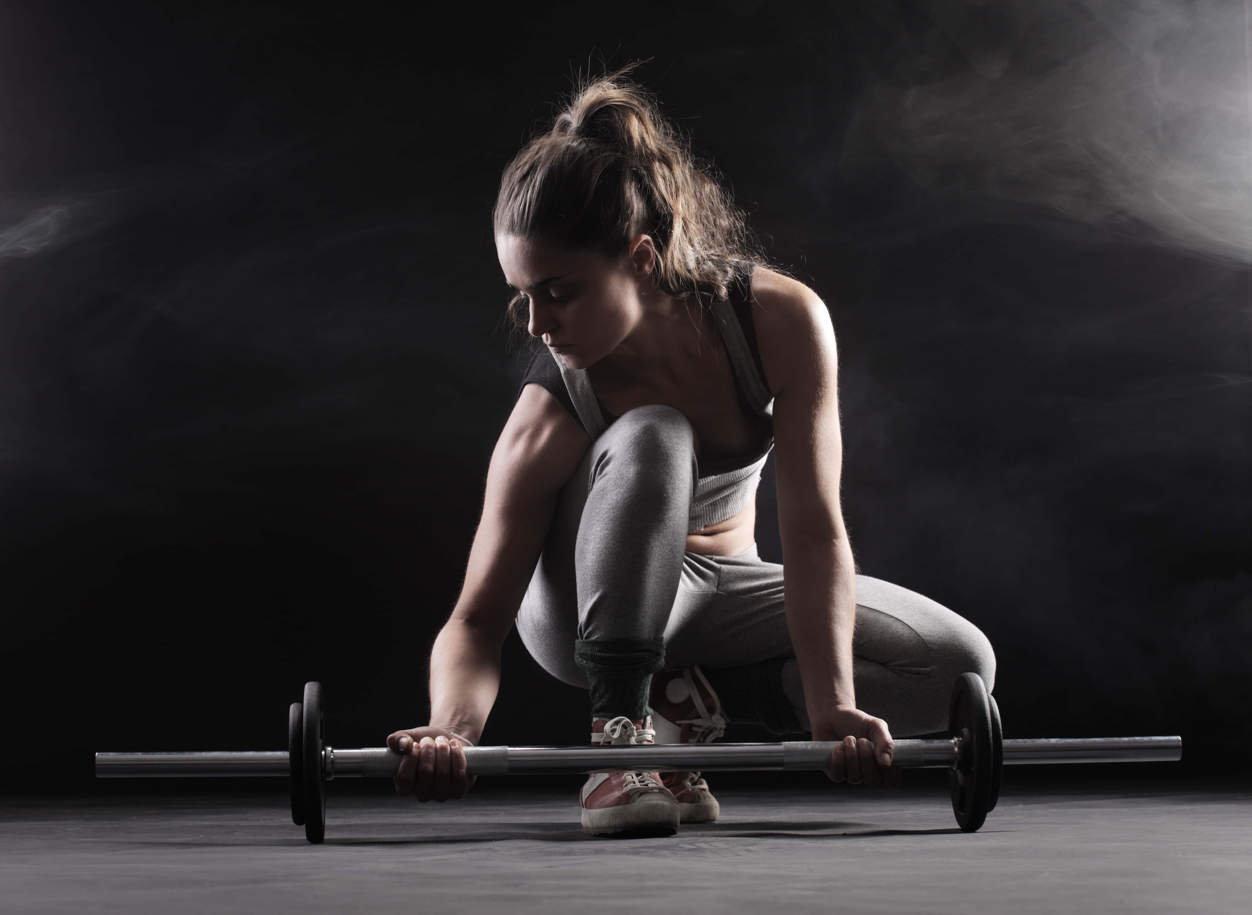 Barbell 桌布 壁紙 — 一位專注的女運動員穿著運動服，在煙霧瀰漫的黑暗工作室背景下低蹲抓握槓鈴。