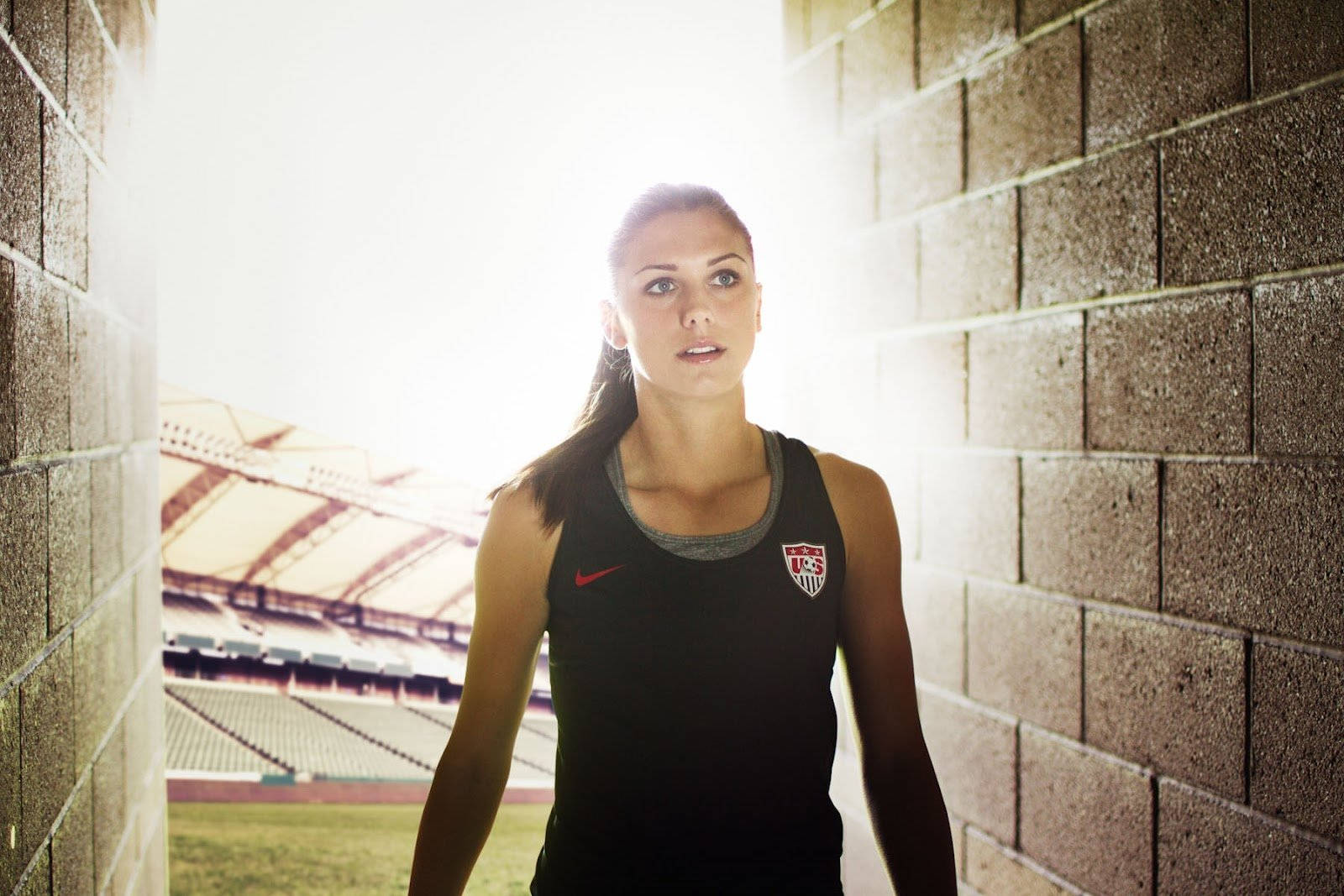 Alex Morgan 桌布 壁紙 — 一位專注的女運動員身穿黑色美國足球球衣，站在陽光普照的體育場隧道中向前看。