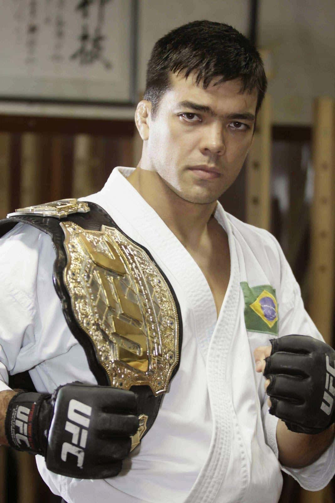 iPhone 桌布 壁紙 — 一位專注的巴西MMA拳手，身穿白色道服，肩上披著金色UFC冠軍腰帶，戴著黑色手套，站在武術道場中。