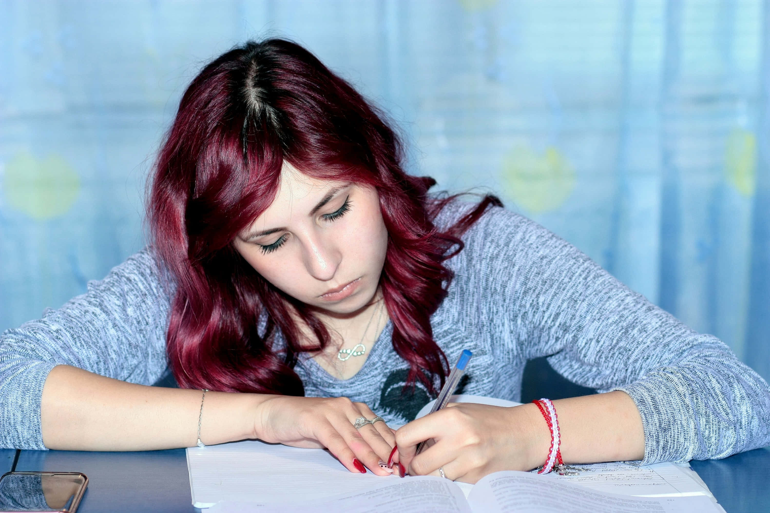 Study Girl 桌布 壁紙 — 一位專注的年輕女性，擁有鮮豔的紅髮，在柔和的藍色窗簾下的書桌前專注地寫筆記。