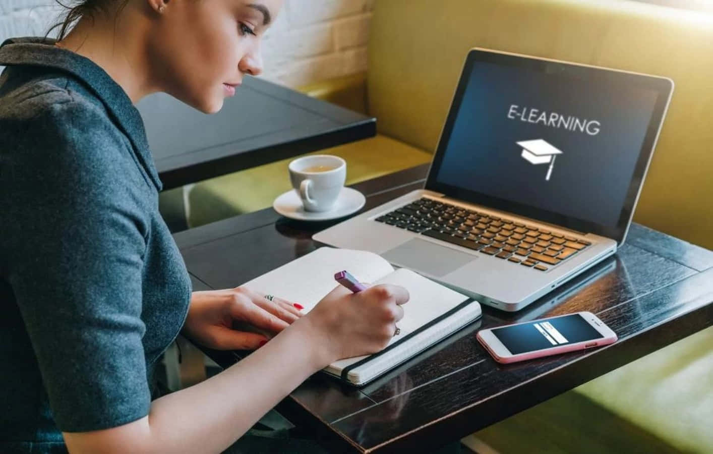Learning 桌布 壁紙 — 一位專注的年輕女性在咖啡館裡，旁邊放著一杯咖啡，一邊在筆記型電腦上學習電子學習內容，一邊寫筆記。