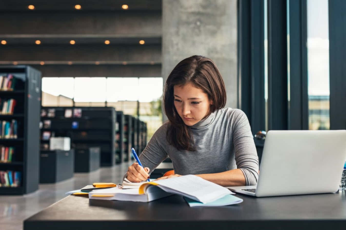 Study Girl 桌布 壁紙 — 一位專注的年輕女性在圖書館桌上，在打開的筆記型電腦旁，於柔和自然光下寫筆記。
