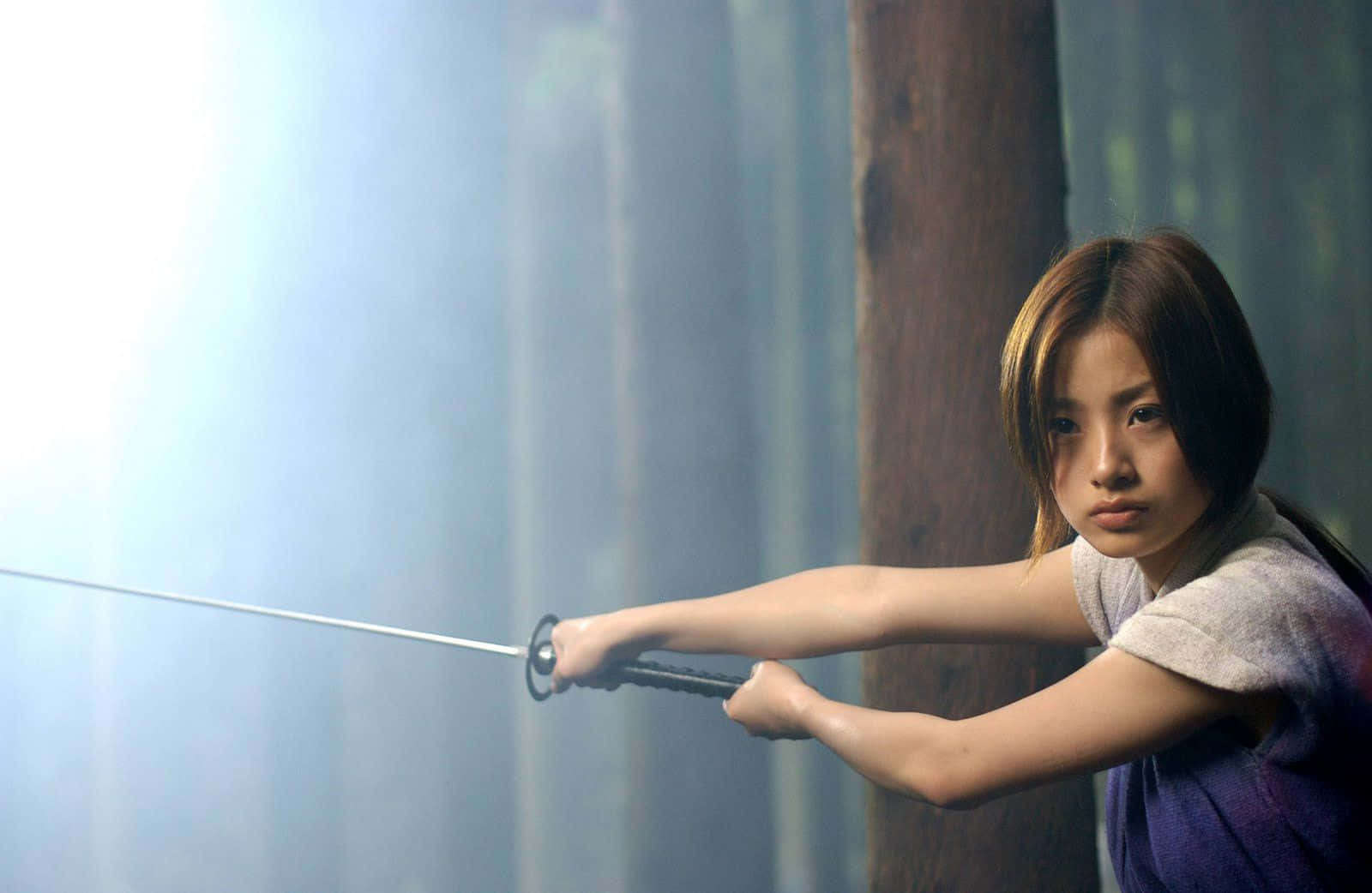Samurai Movies 桌布 壁紙 — 一位專注的年輕女性在森林中，陽光穿過霧氣繚繞的樹木，她帶著強烈的決心握著一把刀。