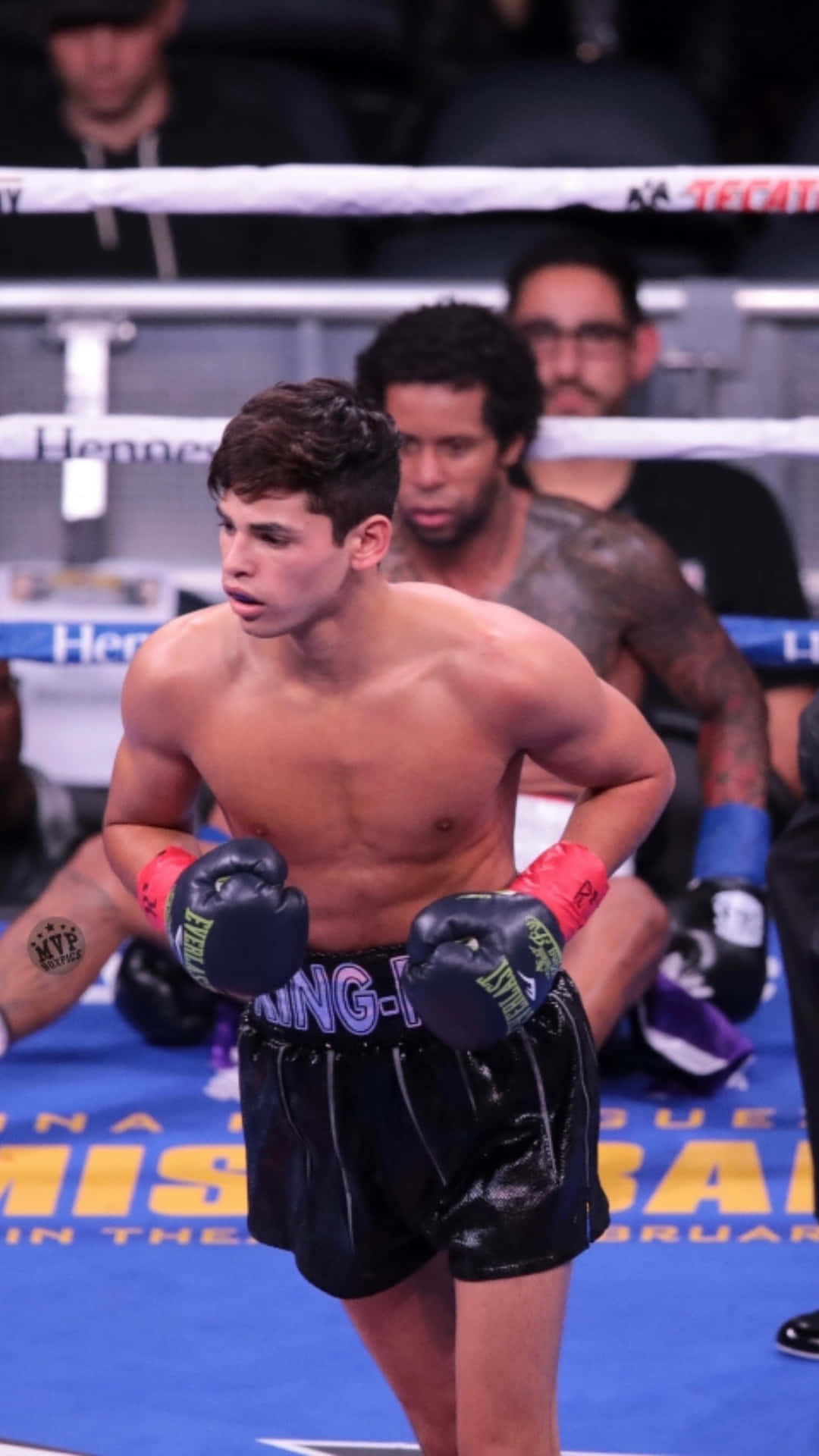 Ryan Garcia 桌布 壁紙 — 一位專注的年輕拳擊手，身穿黑色短褲和手套，在燈光明亮的擂台上警覺地站立，背景有觀眾。