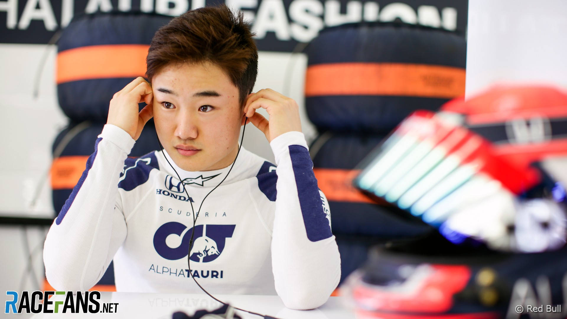 yuki tsunoda 桌布 壁紙 — 一位專注的年輕賽車手穿著AlphaTauri車隊服裝，在賽車活動中調整耳機。