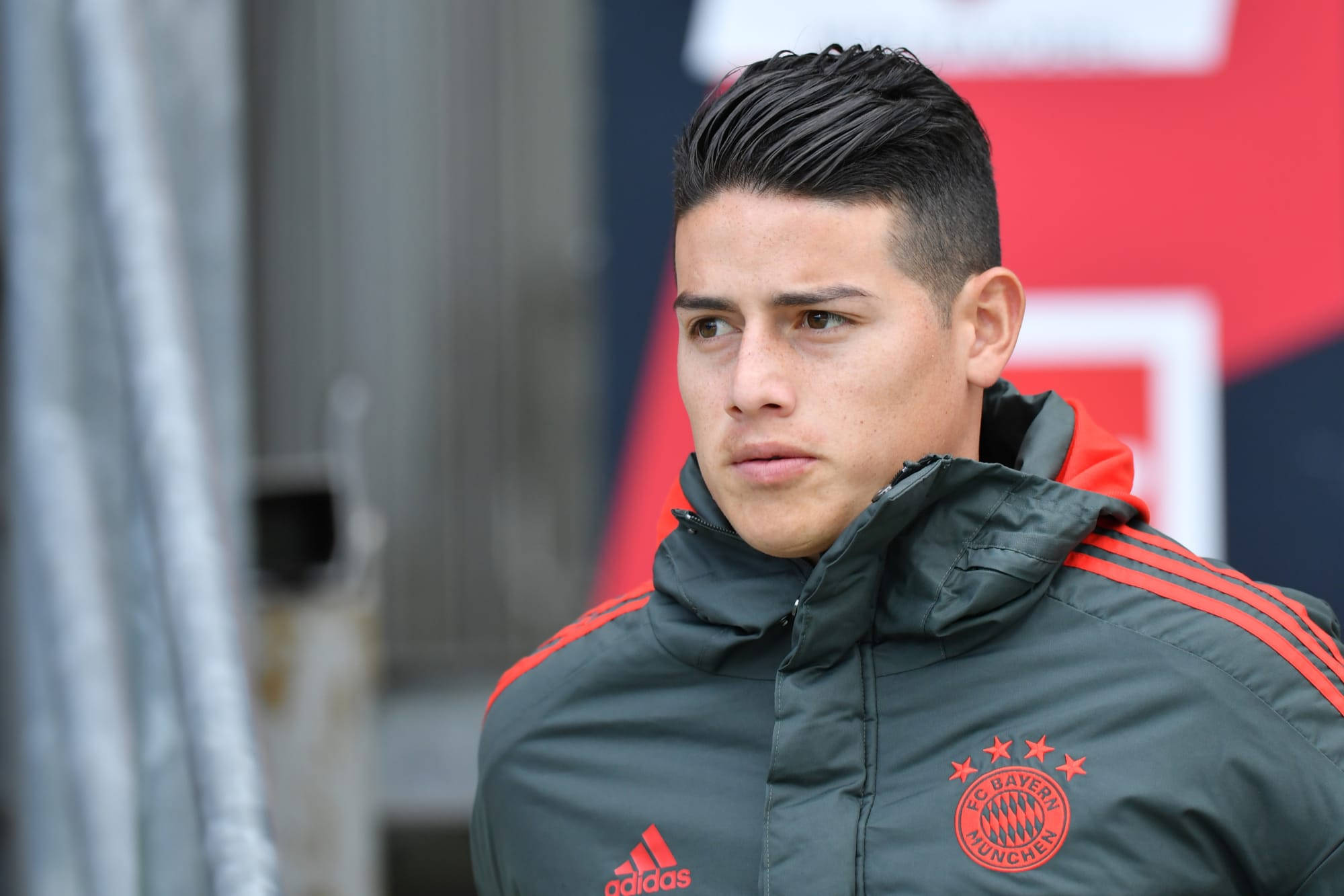 James Rodriguez 桌布 壁紙 — 一位專注的年輕足球員穿著拜仁慕尼黑訓練外套，在模糊的球場背景下凝視鏡頭外。