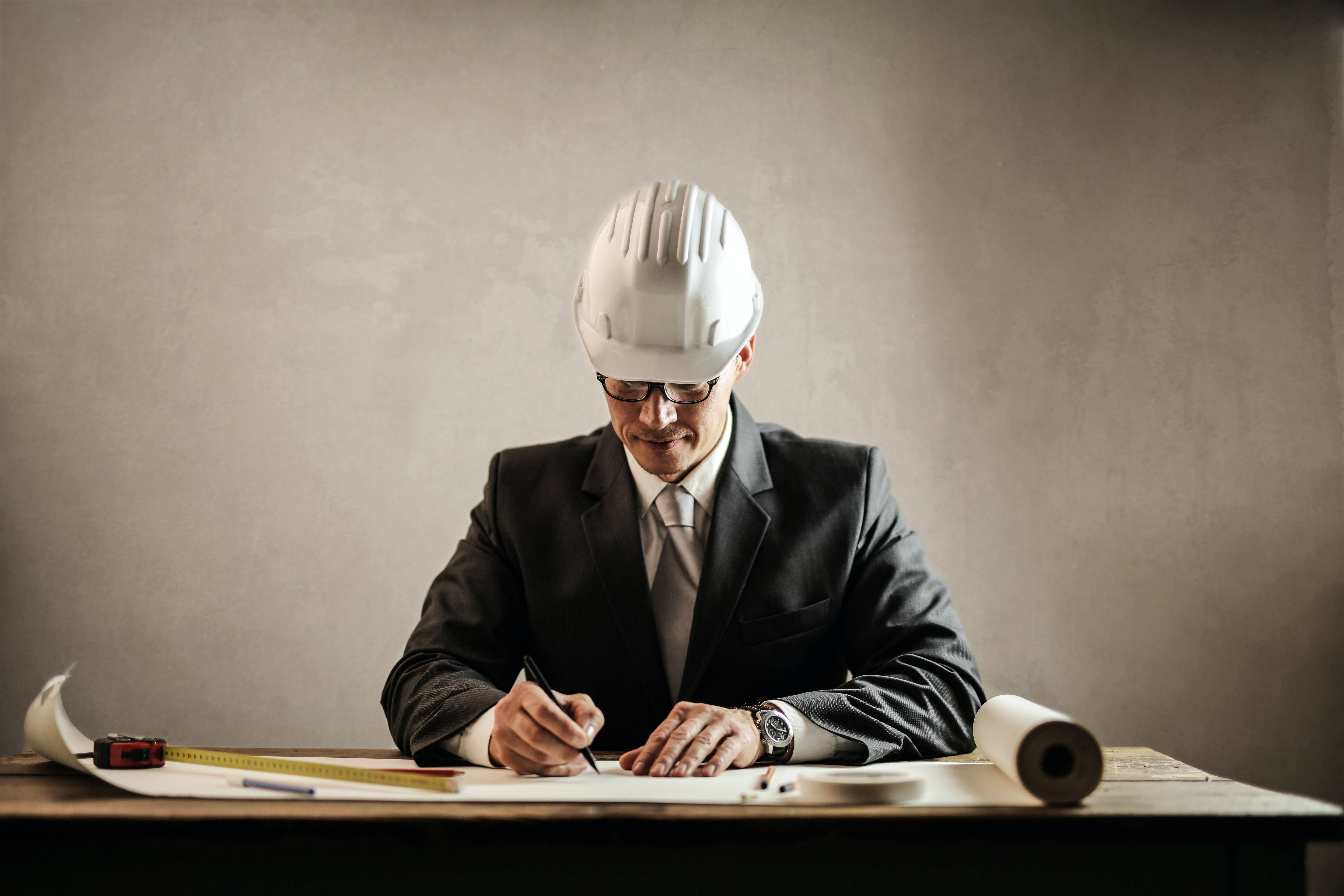 job 桌布 壁紙 — 一位專注的建築師，戴著白色頭盔、穿著西裝，在木桌上簽署藍圖，桌上有工具和捲起的計劃。