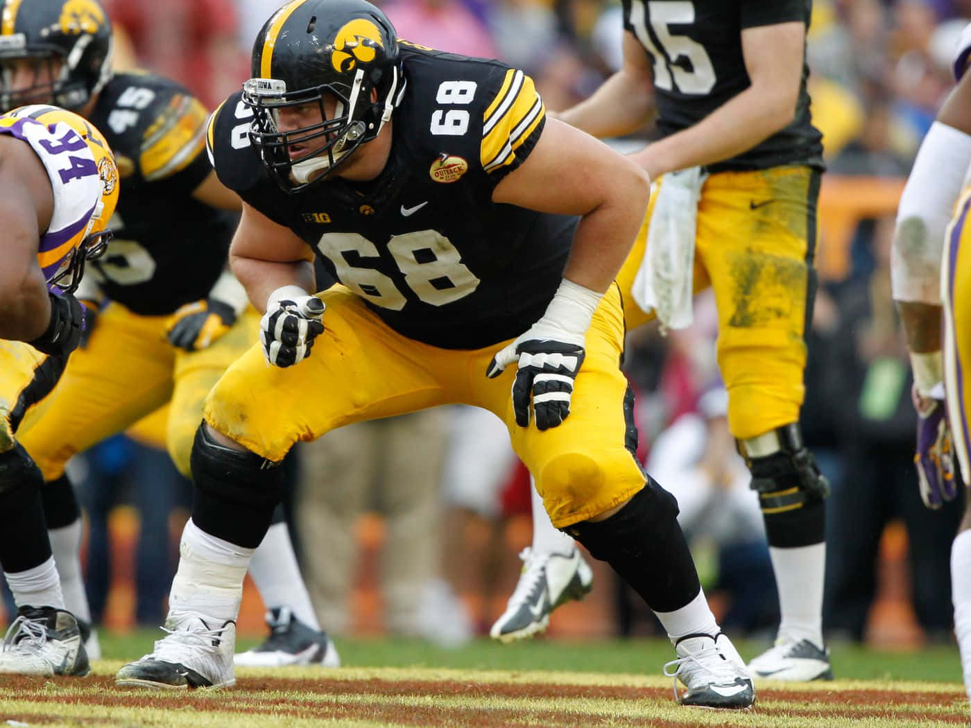 Iowa Hawkeyes 桌布 壁紙 — 一位專注的愛荷華鷹眼隊美式足球員身穿黑色和金色裝備，在球場上低蹲準備行動。