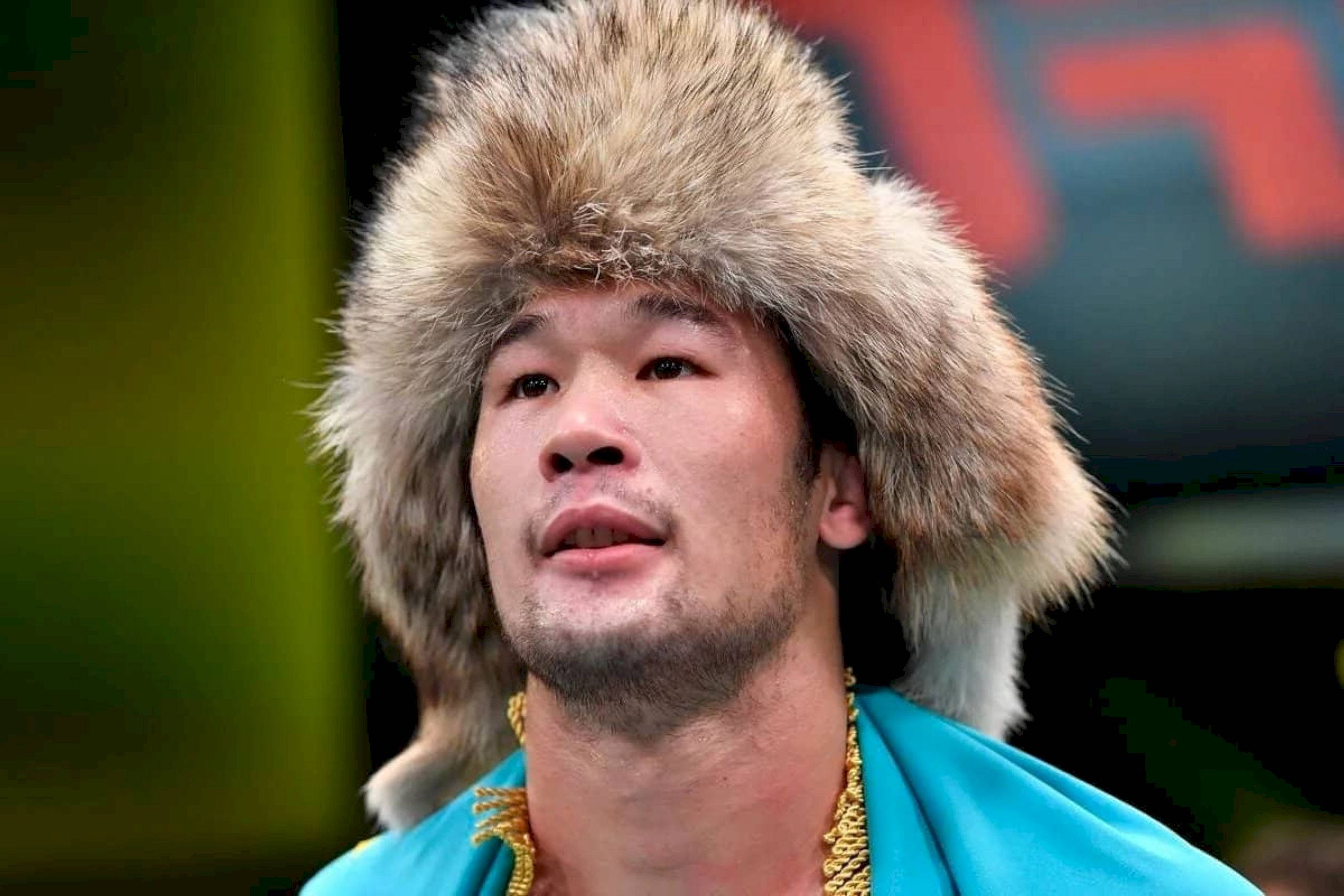 UFC 桌布 壁紙 — 一位專注的戰士戴著毛茸茸的皮帽，身穿帶有金鏈的藍綠色長袍，背景模糊。