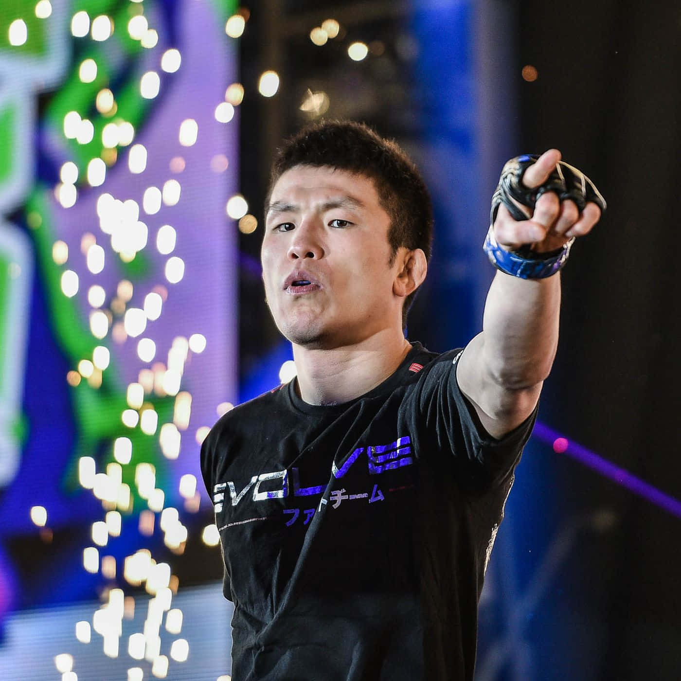 martial arts 桌布 壁紙 — 一位專注的戰士穿著黑色 'EVOLVE' T恤，在鮮豔的舞台燈光下向前指，背景是模糊的彩色散景。