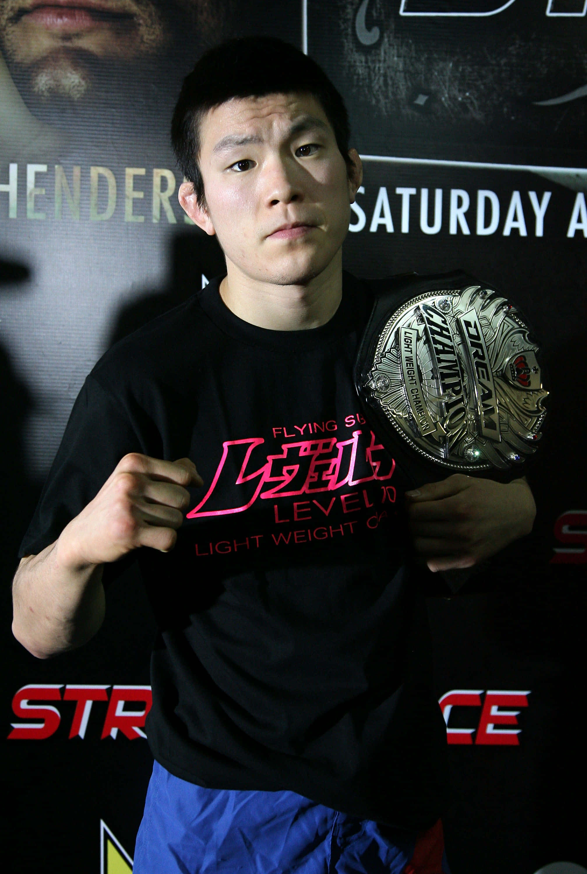 martial arts 桌布 壁紙 — 一位專注的戰士穿著黑色T恤和藍色短褲，在宣傳背景前擺姿勢，手持冠軍腰帶。