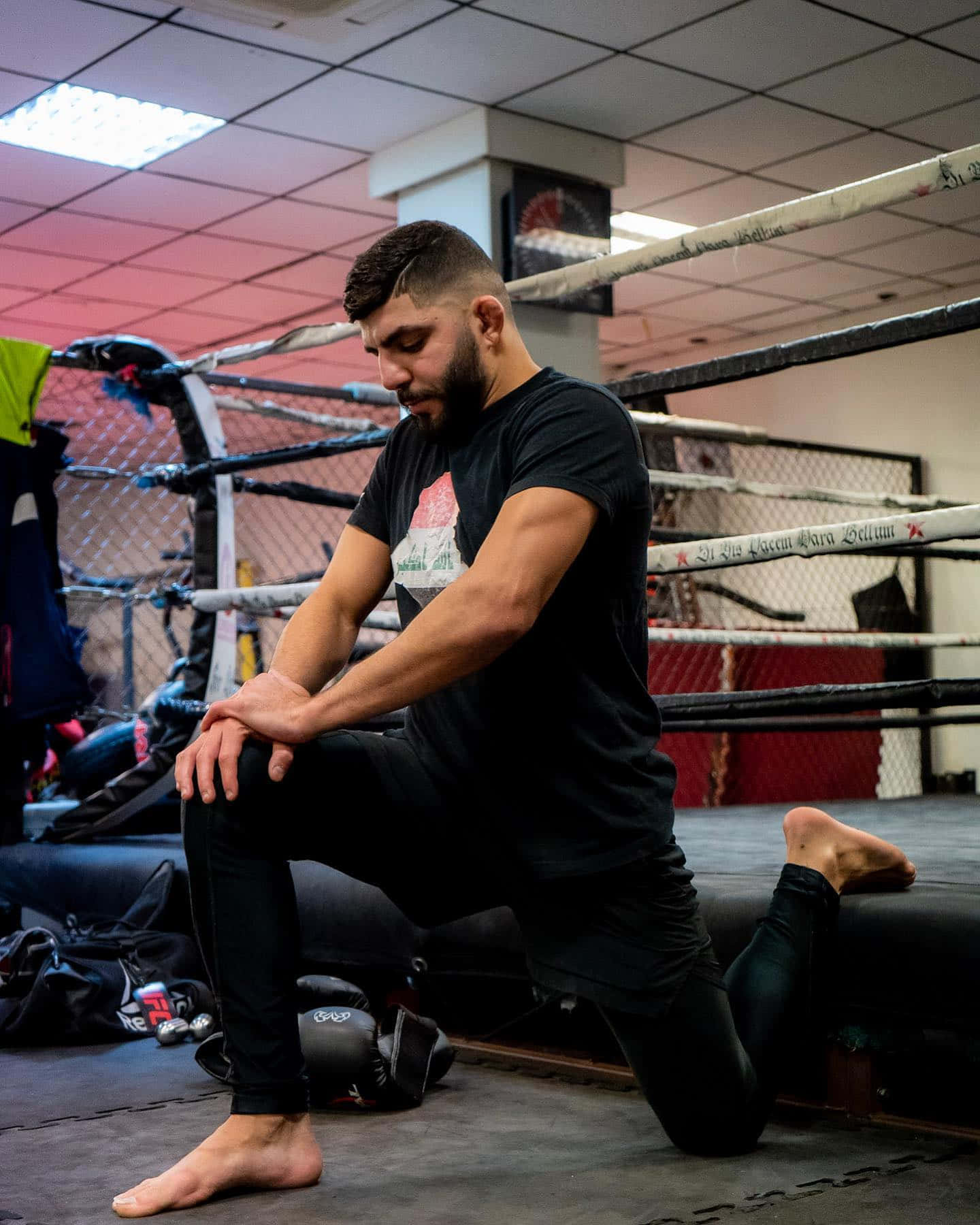 Fitness 桌布 壁紙 — 一位專注的戰士跪在昏暗的MMA訓練擂台內，在訓練前進行伸展。
