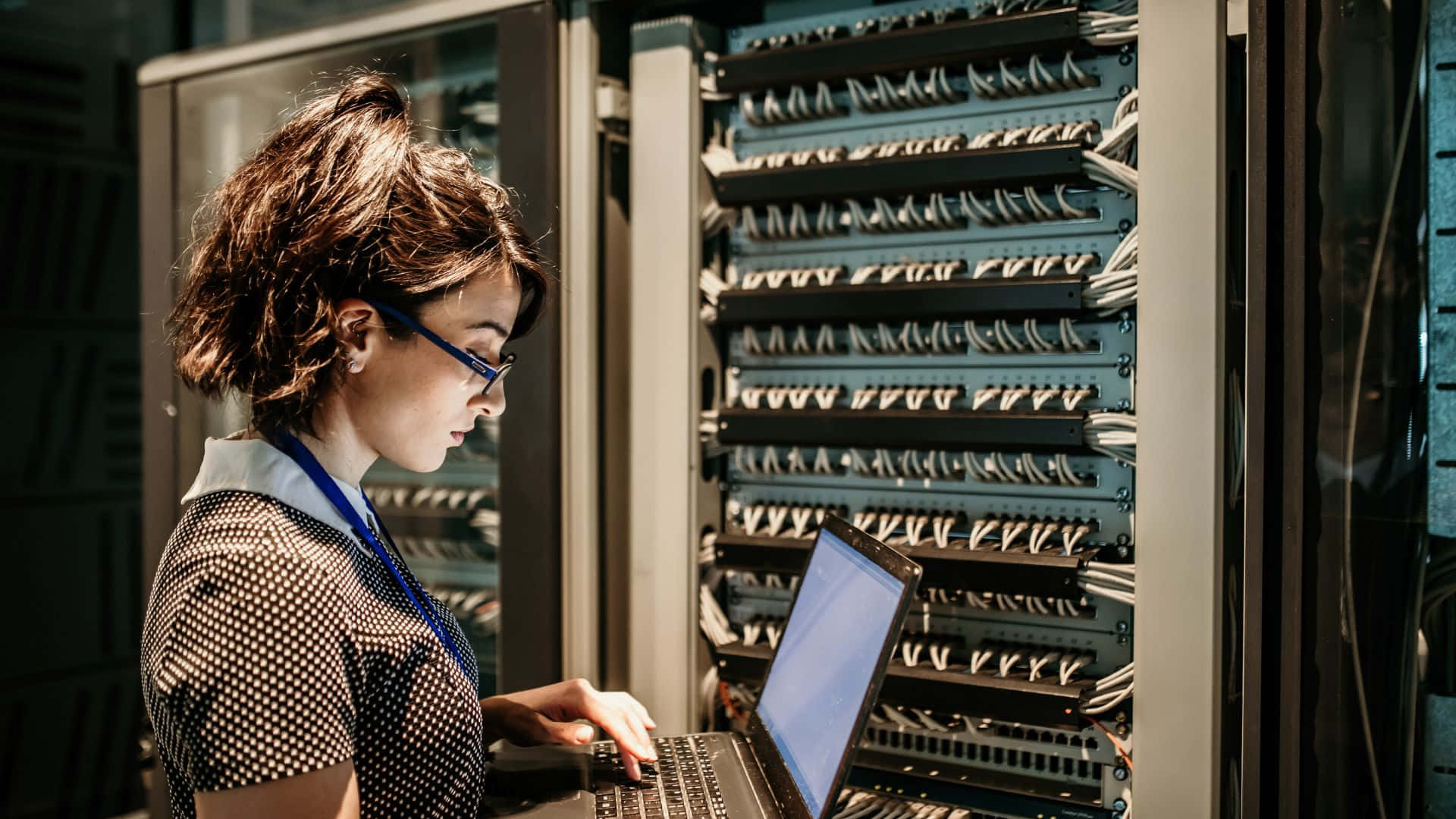 Gaming Servers 桌布 壁紙 — 一位專注的技術人員穿著圓點襯衫，在昏暗的資料中心裡，在 towering 伺服器機架旁邊使用筆記型電腦工作。