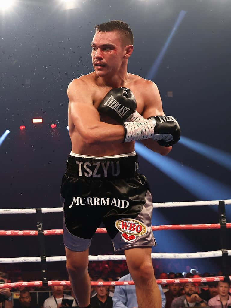 Tim Tszyu 桌布 壁紙 — 一位專注的拳擊手穿著印有「TSZYU」和「MURRMAID」的黑色短褲，在聚光燈下明亮的拳擊場中警覺地站立。