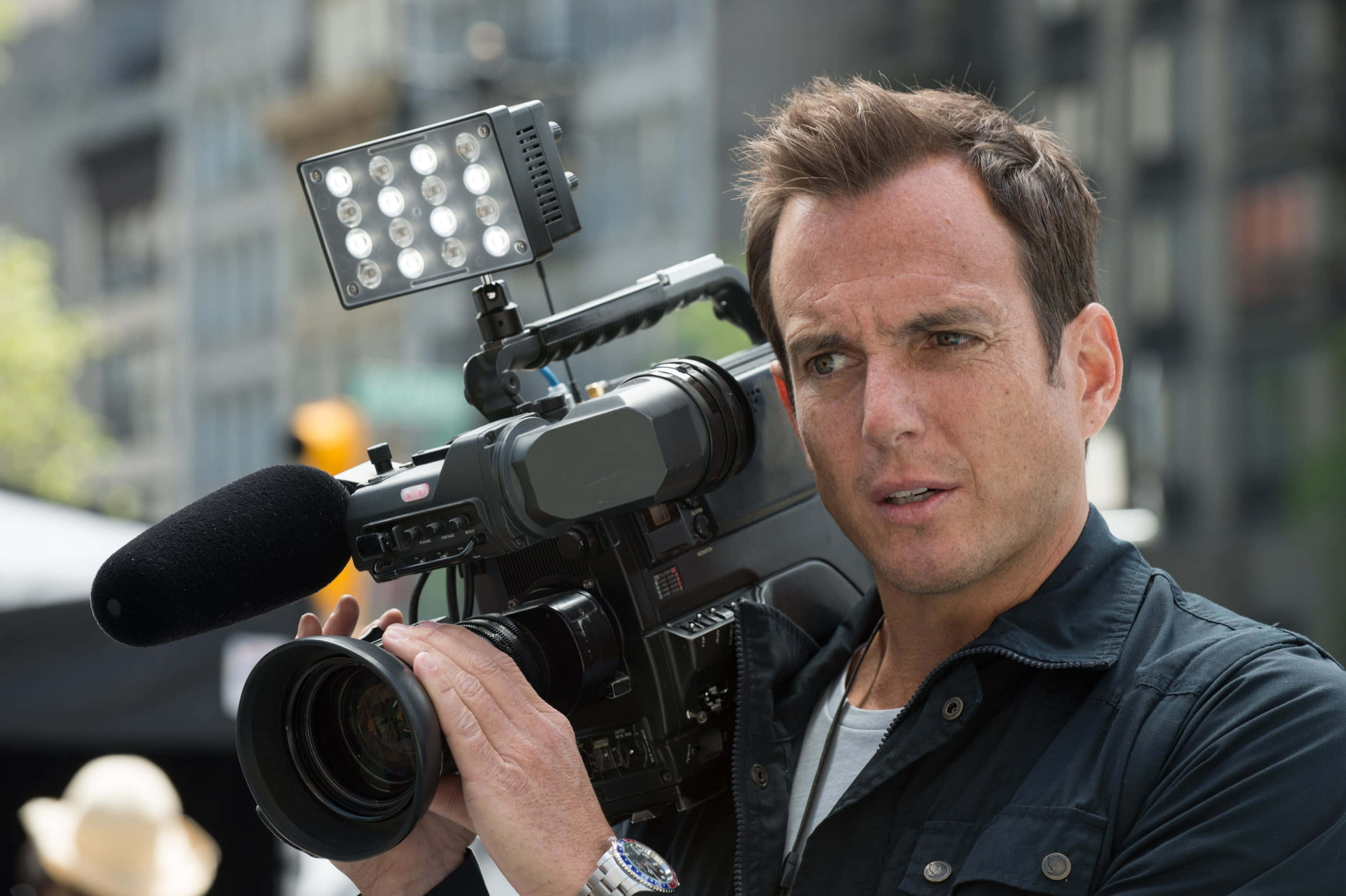 Will Arnett 桌布 壁紙 — 一位專注的攝影師在模糊的城市背景下，手持一部帶有明亮LED燈的專業攝錄機。