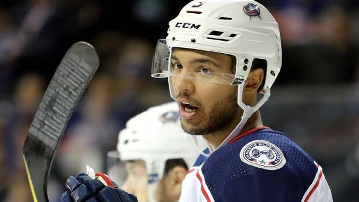 hockey 桌布 壁紙 — 一位專注的曲棍球運動員，戴著白色頭盔和藍色球衣，在比賽中握著球棍，表情專注。
