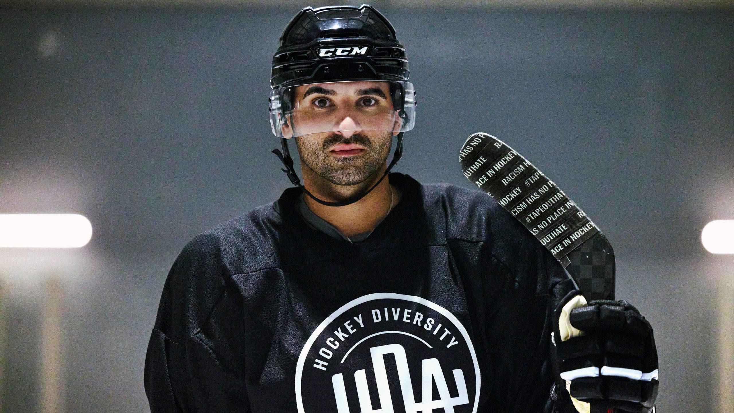 黑色 桌布 壁紙 — 一位專注的曲棍球運動員穿著黑色'Hockey Diversity'球衣，在明亮的場館燈光下握著球桿。