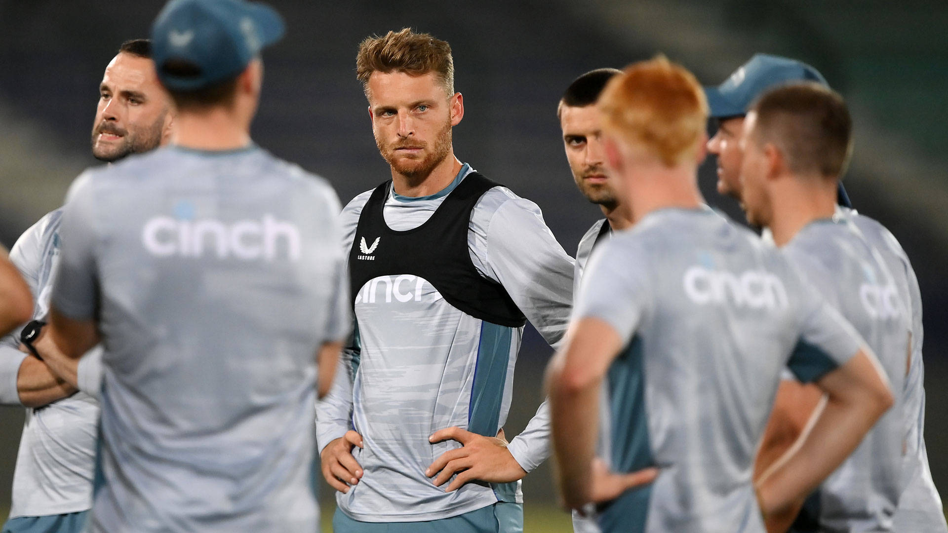 jos buttler 桌布 壁紙 — 一位專注的板球教練穿著黑色背心，在夜間訓練期間於體育場燈光下向他的團隊講話。