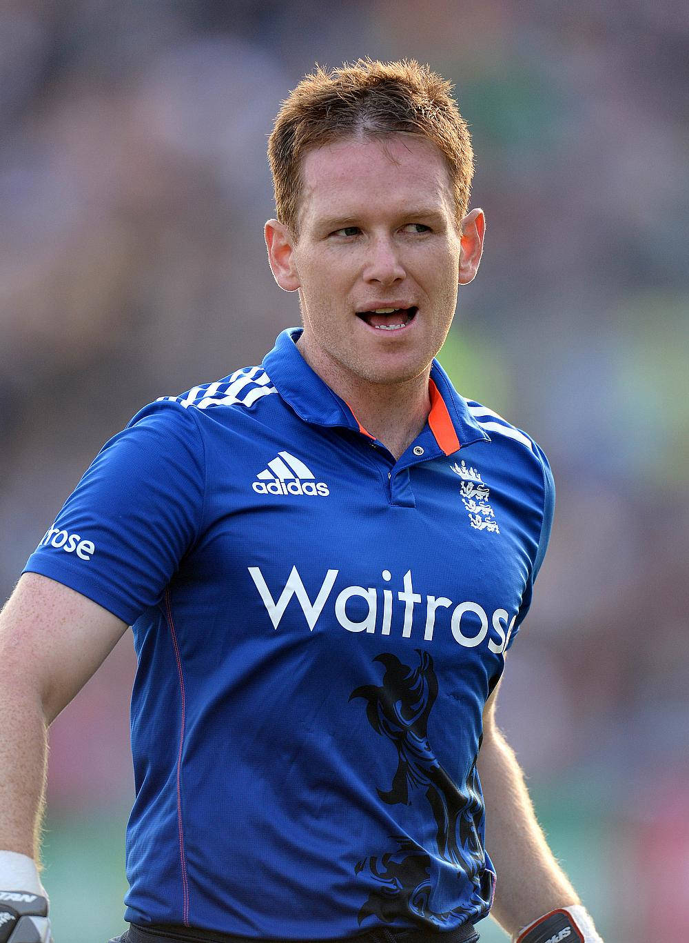 eoin morgan 桌布 壁紙 — 一位專注的板球運動員，身穿帶有Waitrose贊助的藍色英格蘭球衣，站在陽光明媚的球場上。