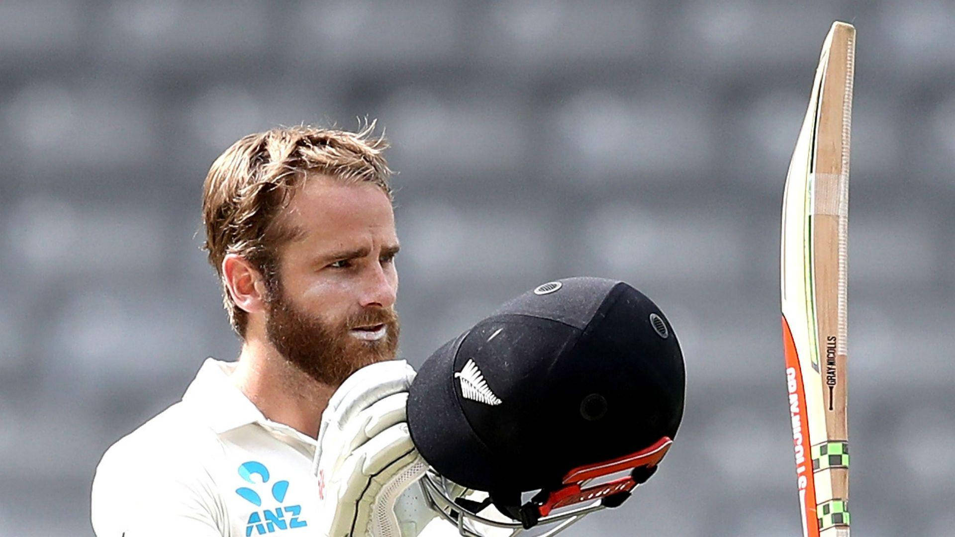 kane williamson 桌布 壁紙 — 一位專注的板球運動員身穿白色制服，在模糊的體育場座位前手持頭盔和球棒。