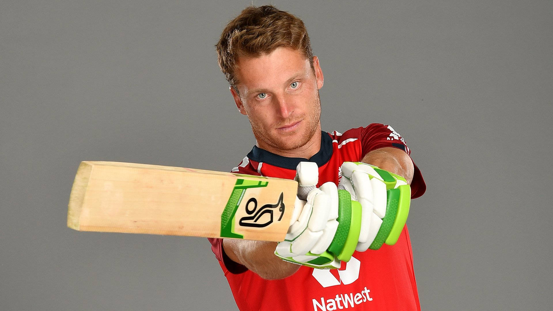 jos buttler 桌布 壁紙 — 一位專注的板球運動員身穿紅色球衣，戴著綠色手套，在純灰色工作室背景前握著木製球棒。