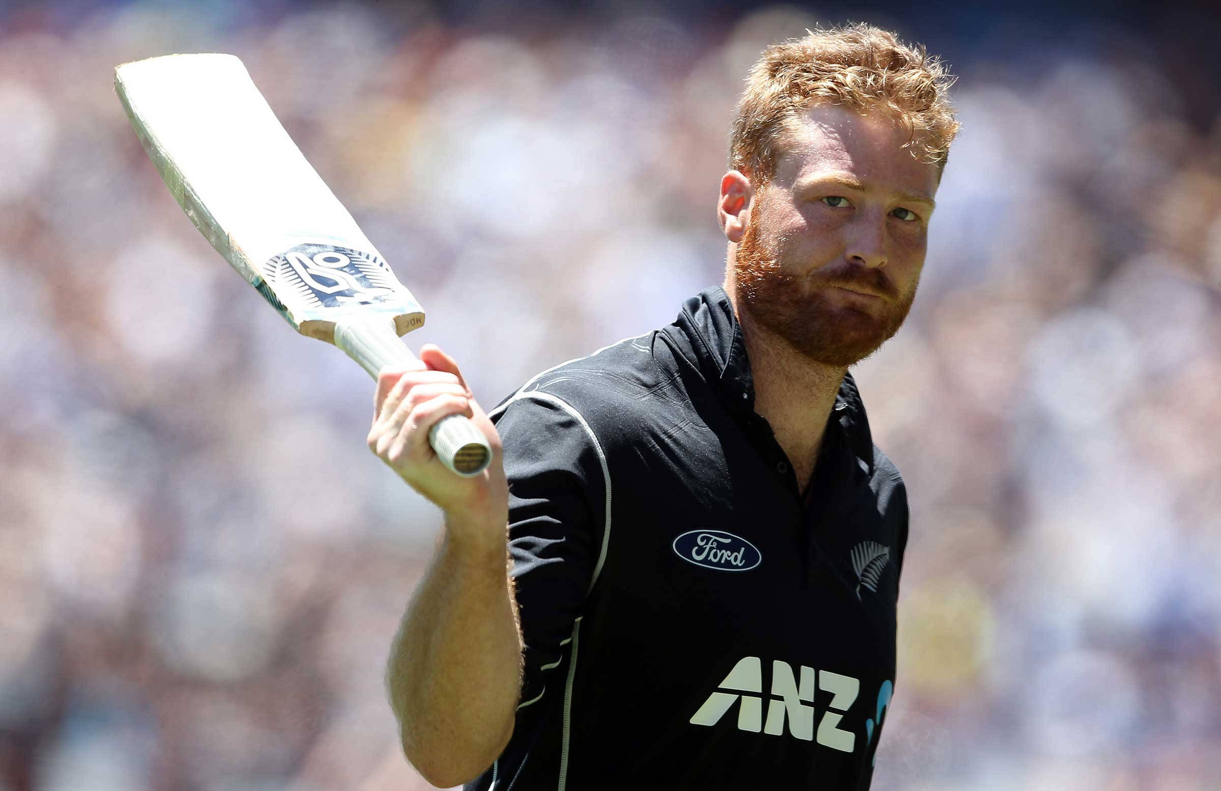 martin guptill 桌布 壁紙 — 一位專注的板球運動員身穿黑色球衣，在明亮的日光下，對著模糊的體育場人群高舉球棒。