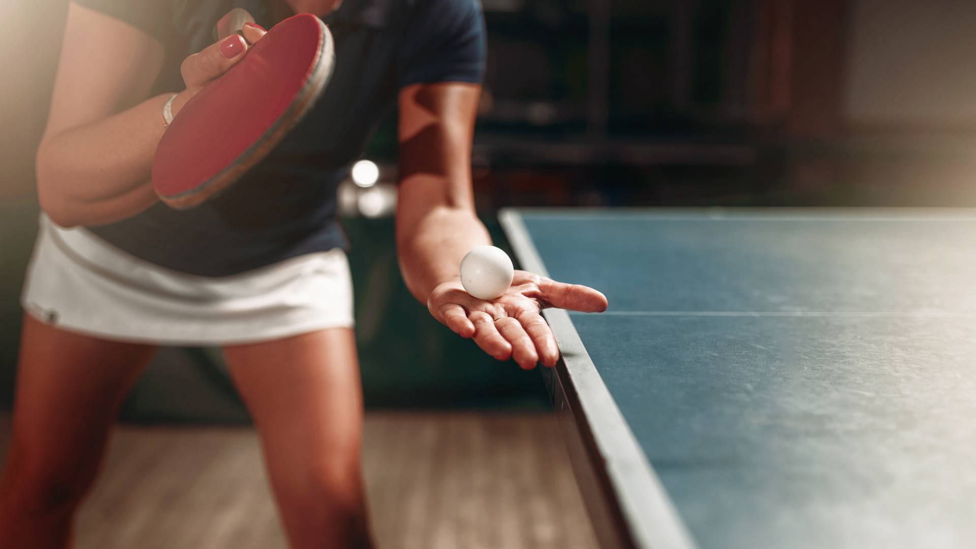 Table Tennis 桌布 壁紙 — 一位專注的桌球運動員在溫暖的室內燈光下，在藍色球桌上拿著球拍和球準備行動。