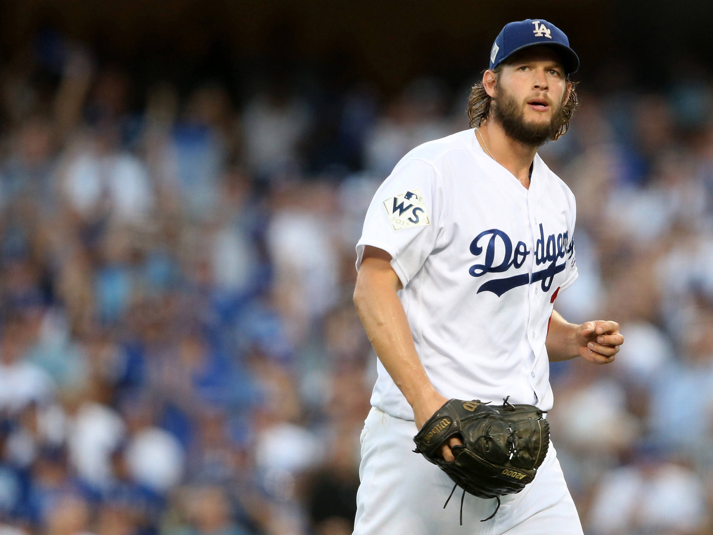 La Dodgers 桌布 壁紙 — 一位專注的洛杉磯道奇隊棒球投手，身穿白色球衣和藍色帽子，站在球場上，身後是模糊的體育館人群。