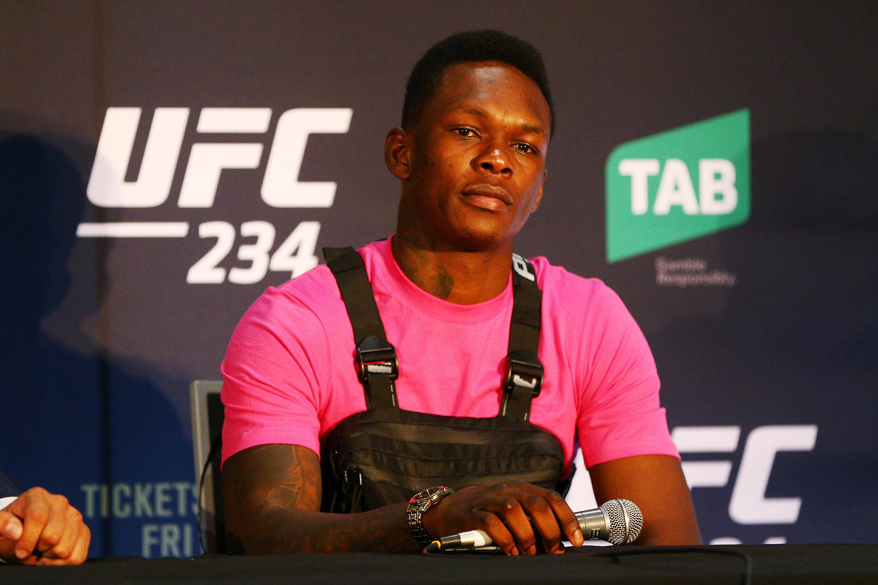 Adesanya 桌布 壁紙 — 一位專注的混合武術家，身穿亮粉色襯衫和黑色背心，坐在記者會桌前，面前有麥克風。