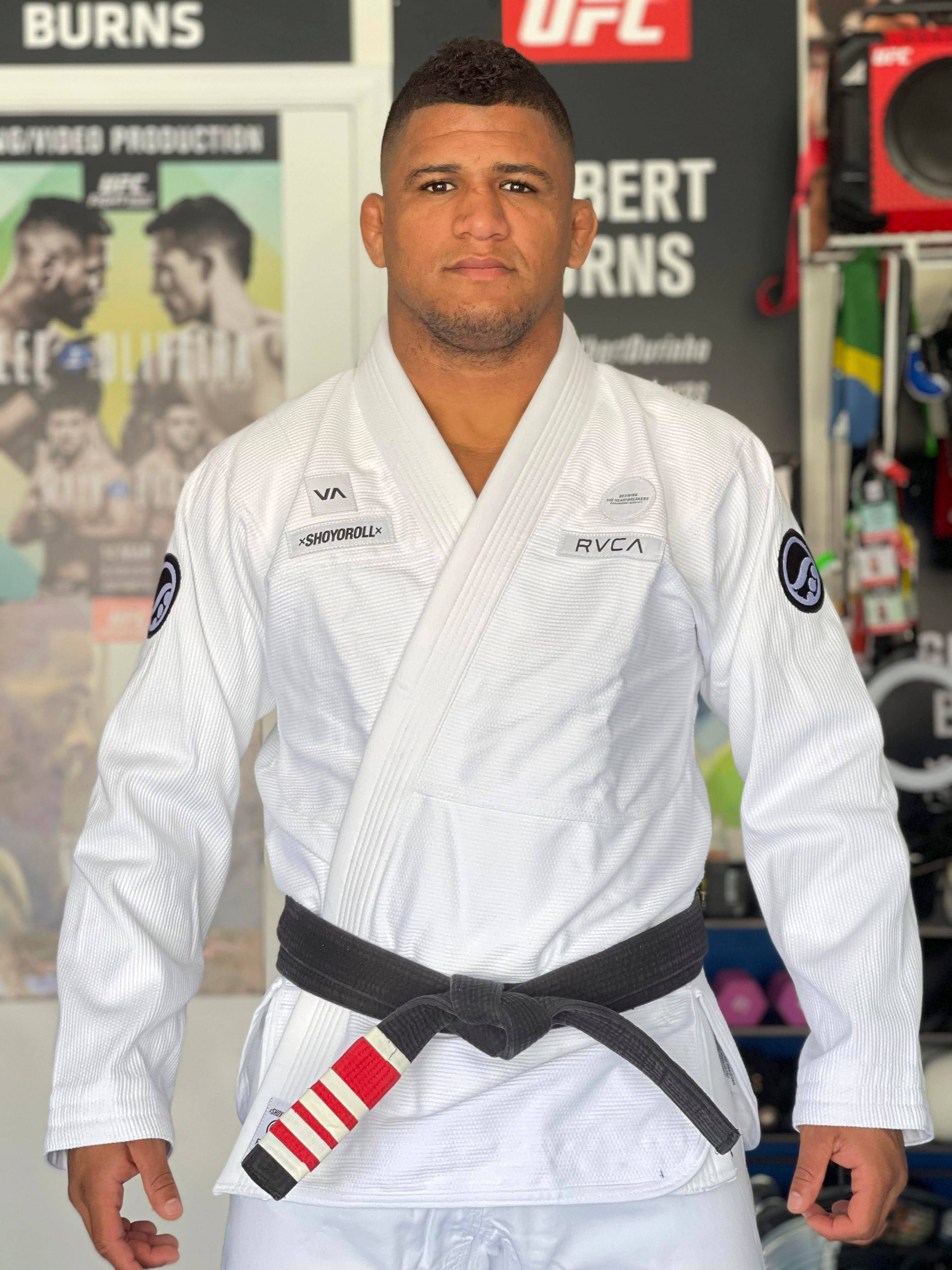MMA 桌布 壁紙 — 一位專注的混合武術家，身穿白色道服配黑帶，自信地站在裝飾有UFC海報的健身房中。