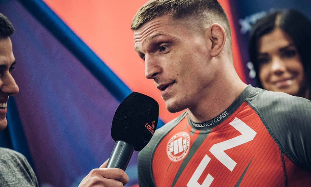 UFC 桌布 壁紙 — 一位專注的混合武術家，身穿紅灰色道服，在賽後採訪中對著麥克風說話，背景人物模糊。