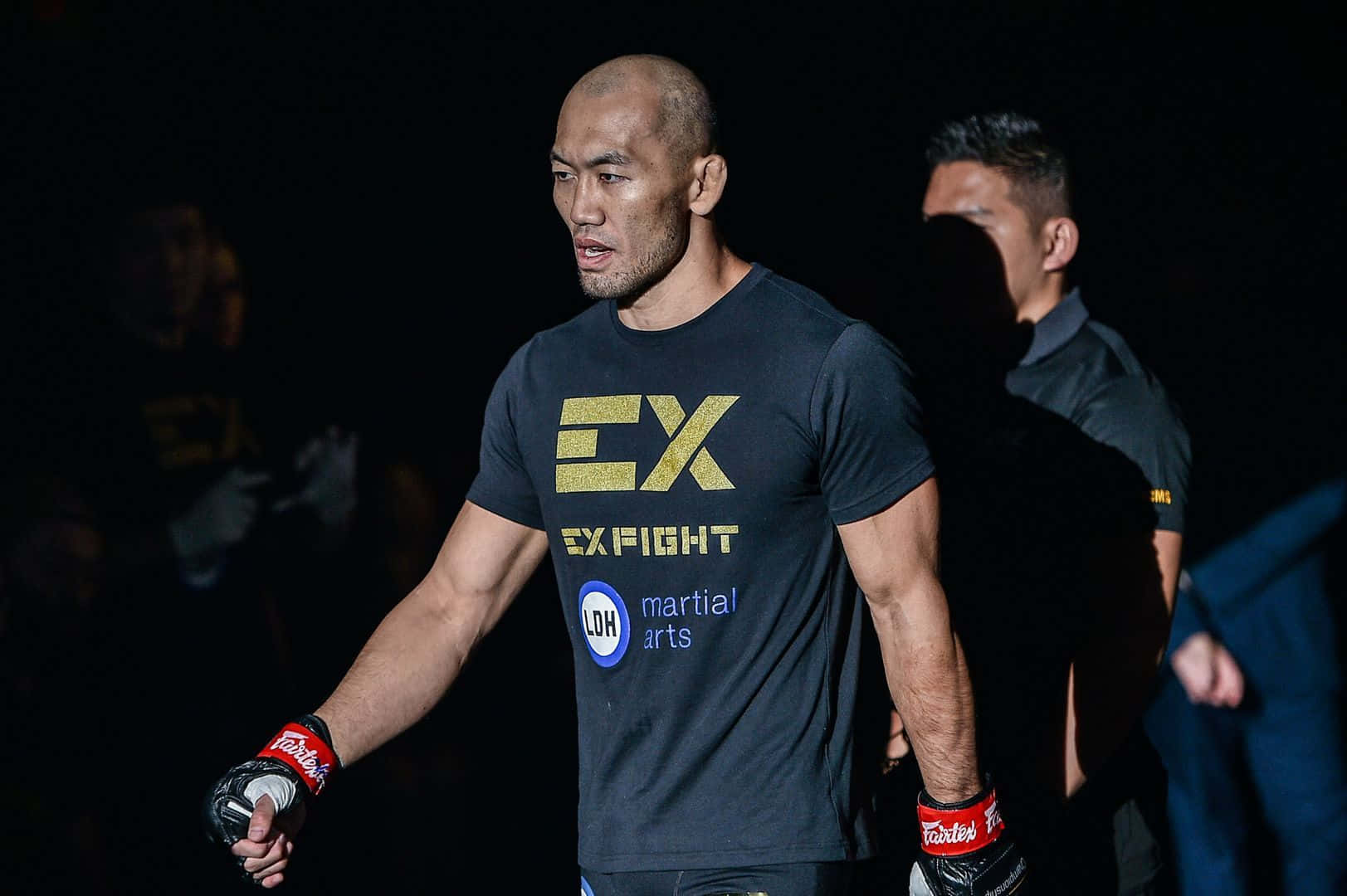 UFC 桌布 壁紙 — 一位專注的混合武術家，身穿黑色裝備，帶有金色 'EX FIGHT' 標誌，在競技場燈光下大步向前。