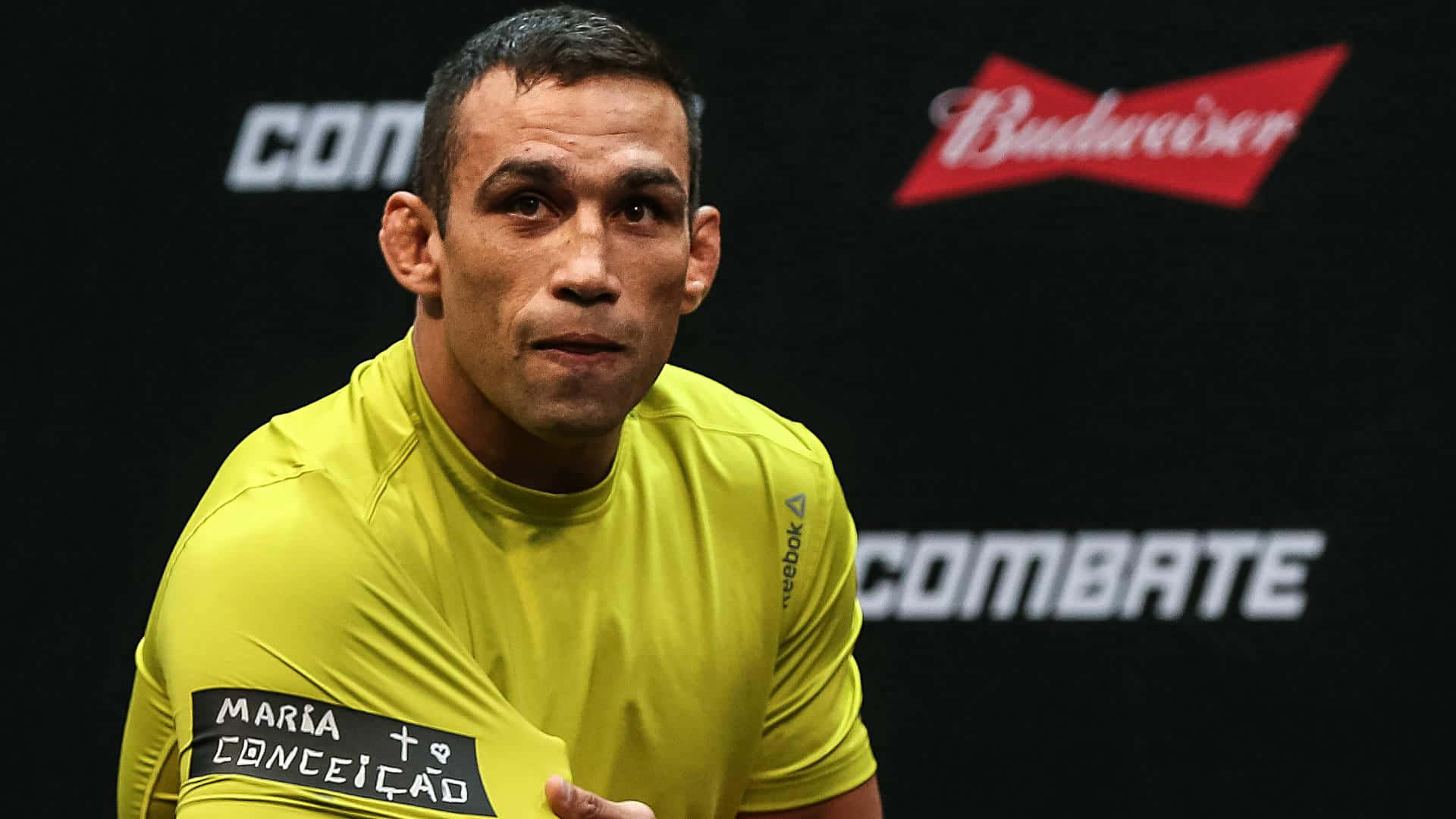 sport 桌布 壁紙 — 一位專注的混合武術家穿著亮黃色 Reebok 襯衫，站在帶有 Budweiser 和 Combate 標誌的黑色背景前。