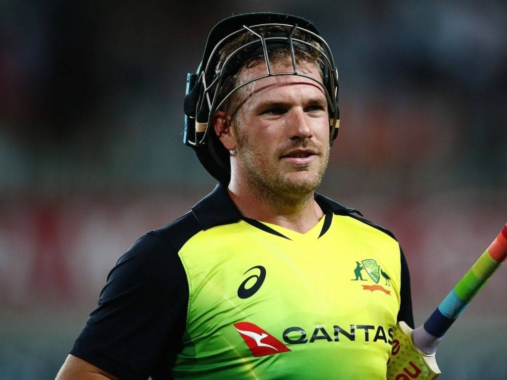 Aaron Finch 桌布 壁紙 — 一位專注的澳洲板球運動員，身穿亮黃綠色球衣和頭盔，在體育館燈光下的晚間比賽中握著球棒。