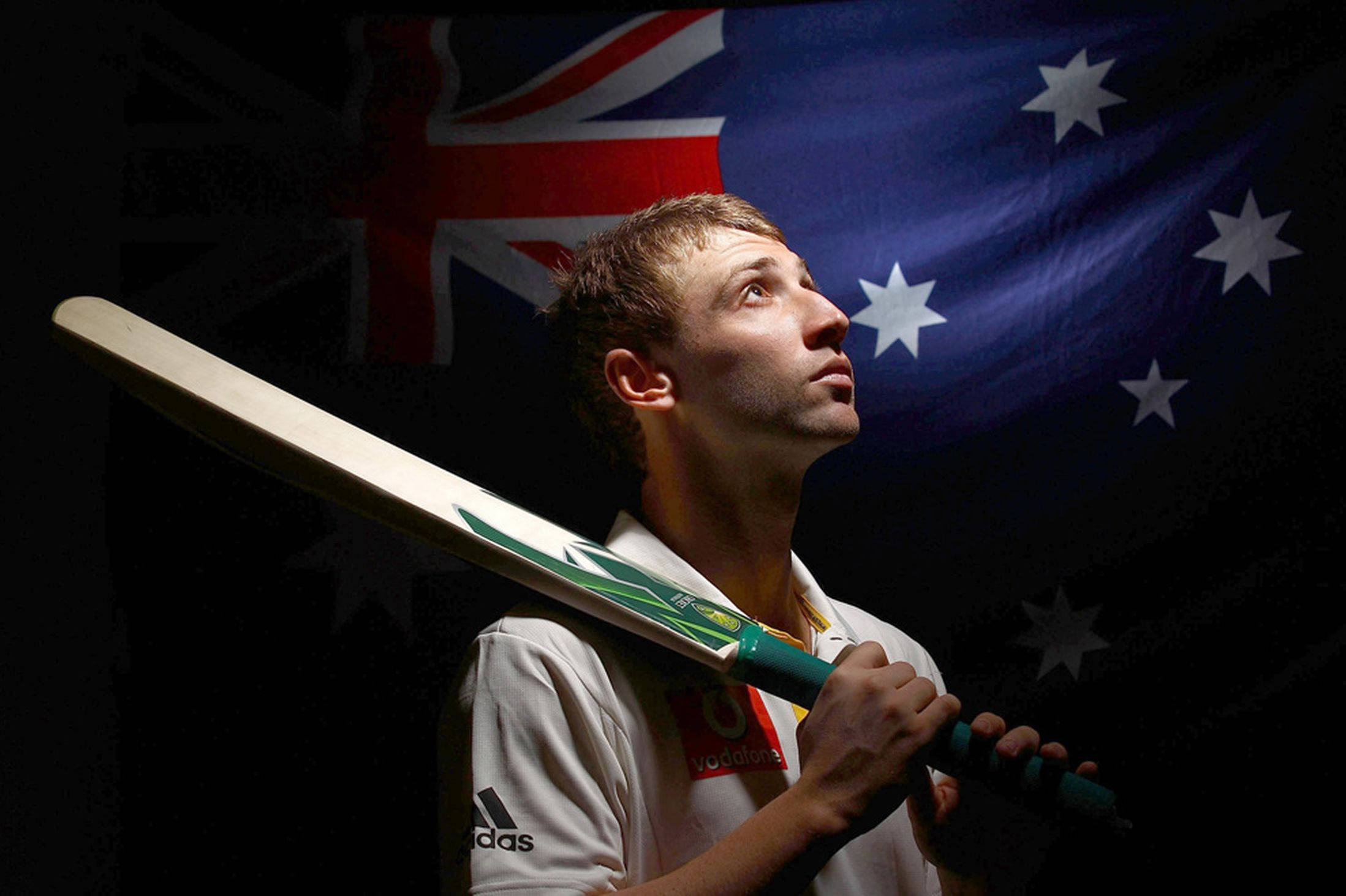 australia cricket 桌布 壁紙 — 一位專注的澳洲板球運動員在黑暗背景下高舉球棒，身後國旗閃耀。