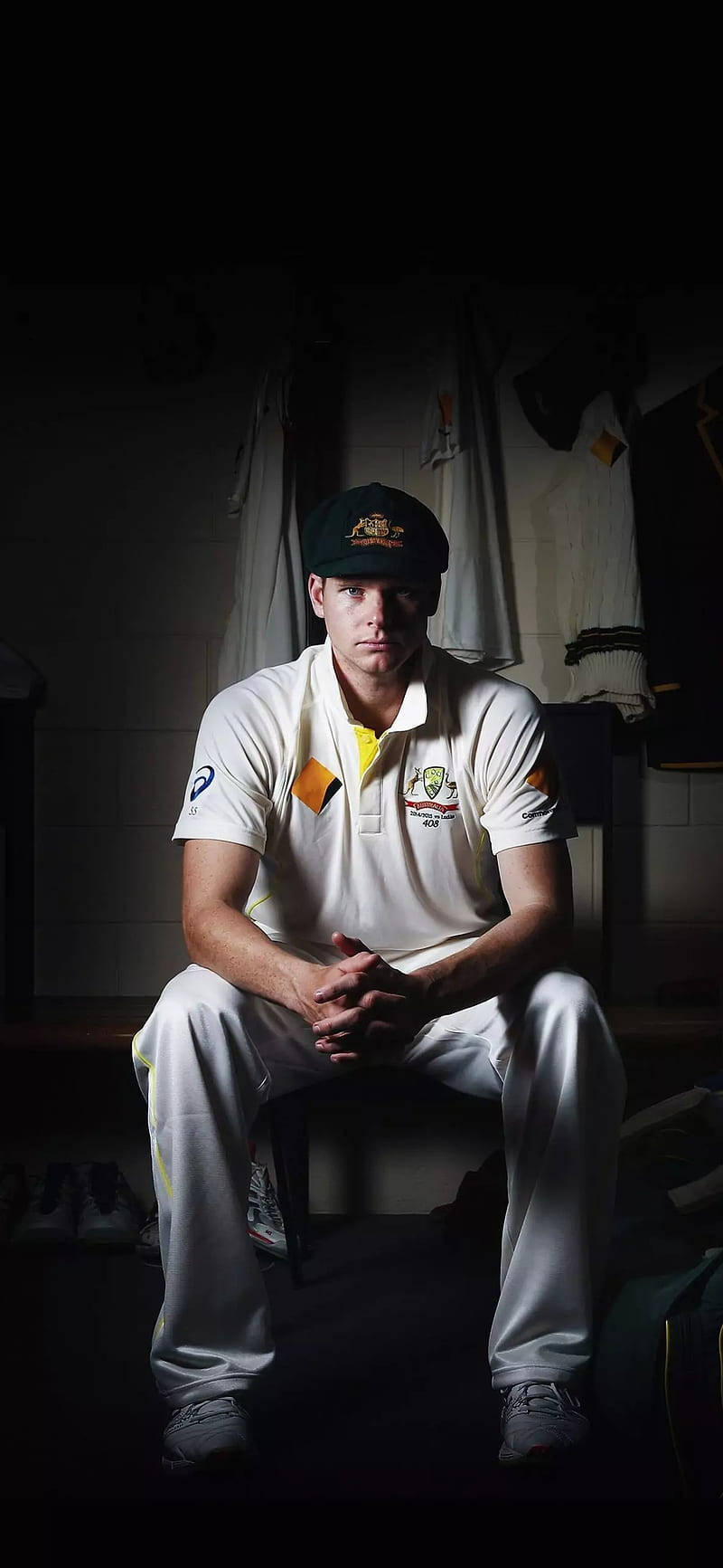 australia cricket 桌布 壁紙 — 一位專注的澳洲板球運動員坐在光線昏暗的更衣室裡，穿著隊服和帽子。