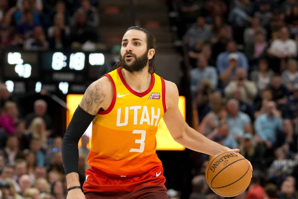 Ricky Rubio 桌布 壁紙 — 一位專注的猶他爵士籃球運動員身穿橙色球衣，在明亮的場館燈光下於激烈的比賽中運球。