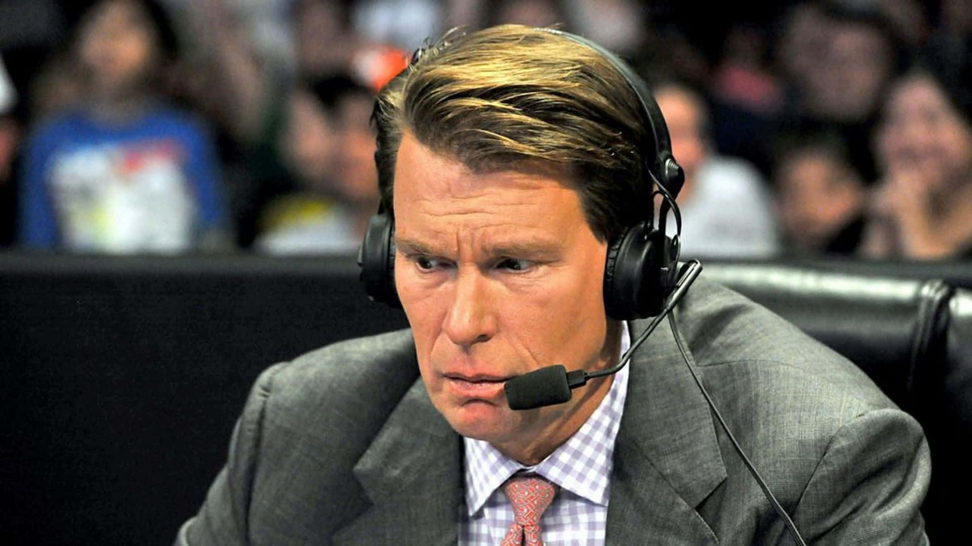 WWE 桌布 壁紙 — 一位專注的男子身穿西裝和耳機，坐在體育轉播室內，身後是模糊的人群。
