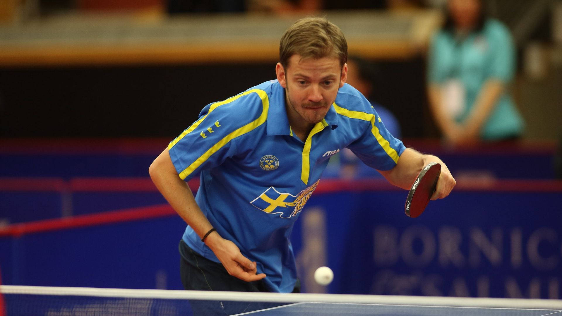 Table Tennis 桌布 壁紙 — 一位專注的男性乒乓球運動員身穿藍黃色球衣，在室內比賽中撲身回擊。