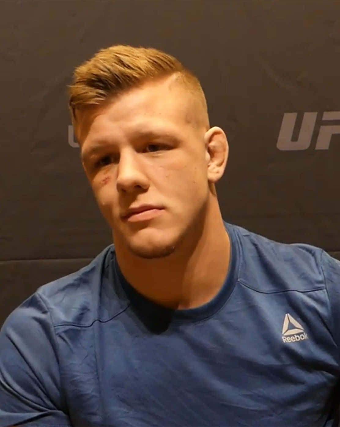 UFC 桌布 壁紙 — 一位專注的男性拳手，臉頰上有褪色的瘀傷，穿著藍色Reebok襯衫，在深色UFC背景下凝視著。